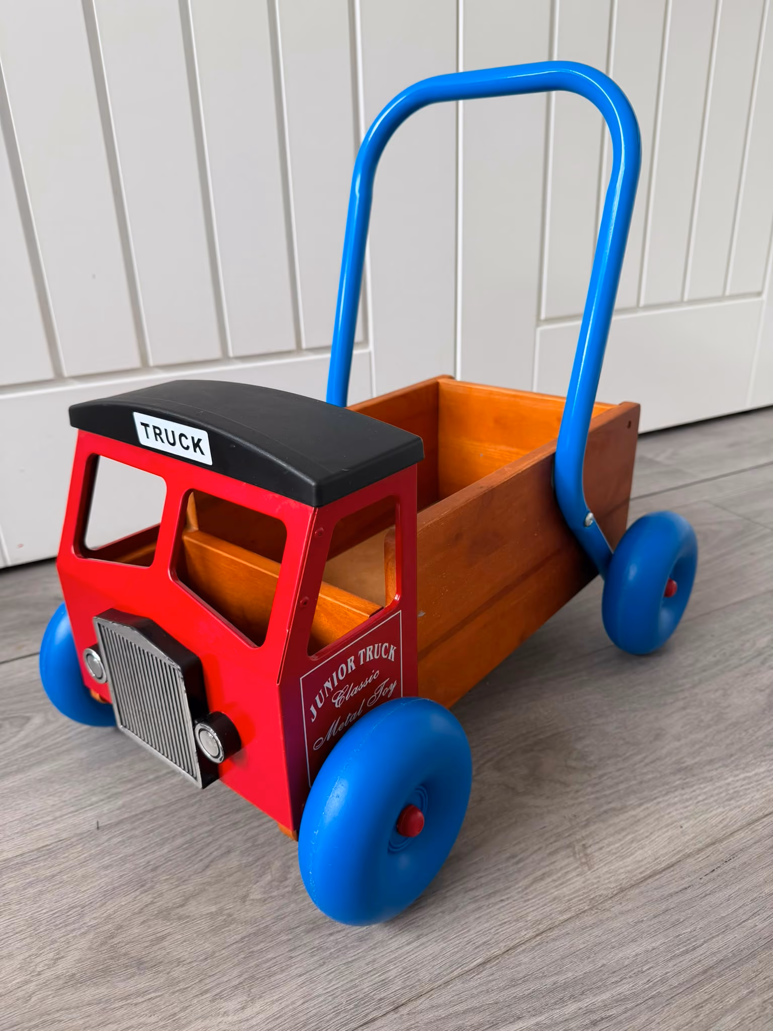 Wooden toy truck with red front, blue wheels and handle, and open cargo bed.