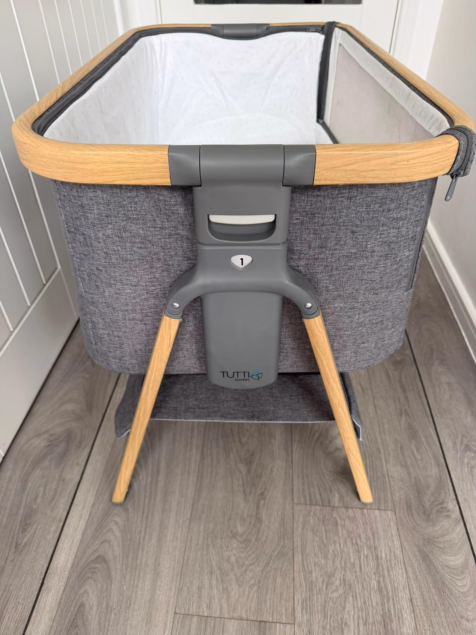 Modern gray baby bassinet with wooden legs and trim on a wood floor inside a room.