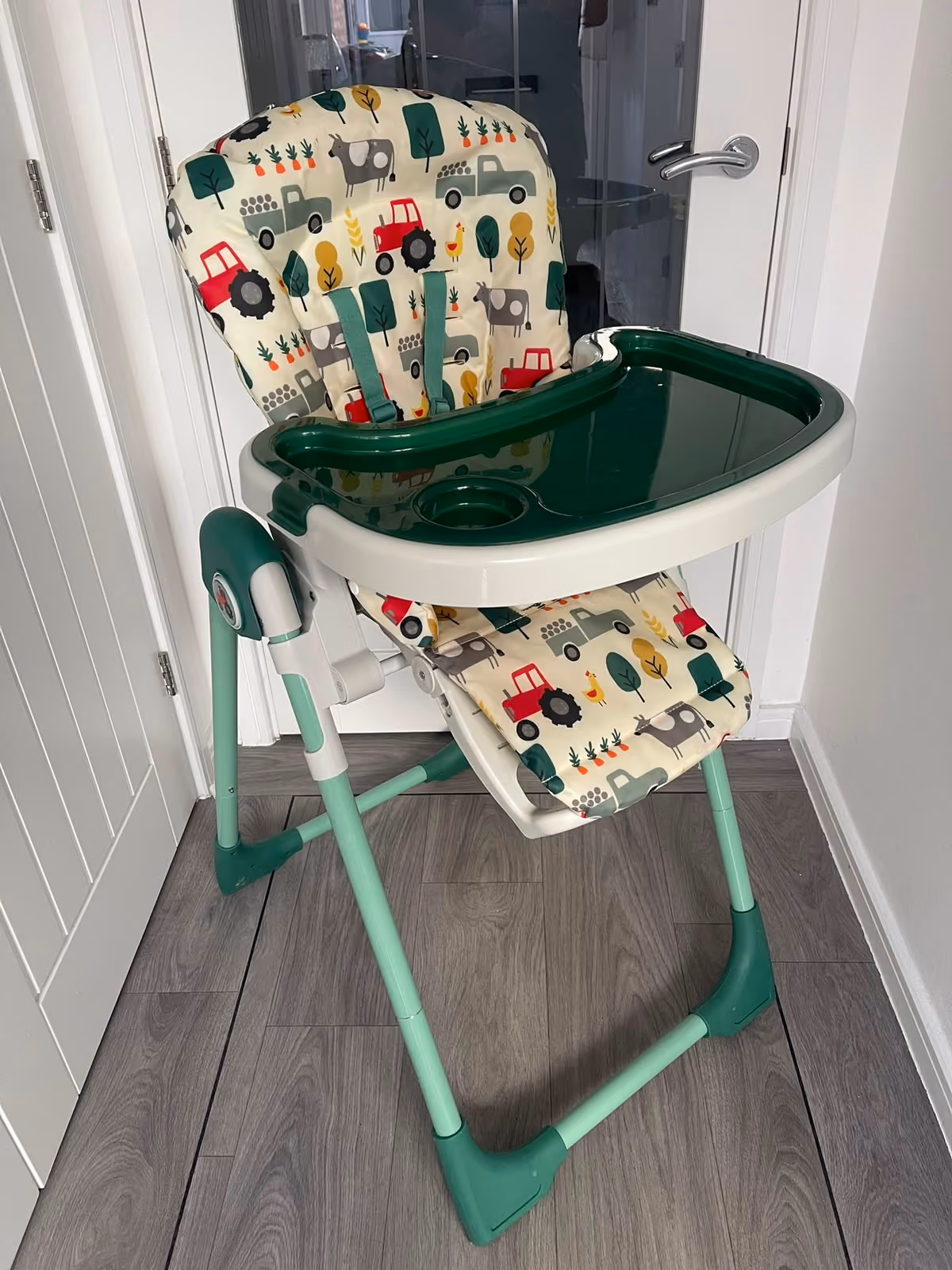 Green and white baby high chair with animal and tractor print cushion on wood flooring.