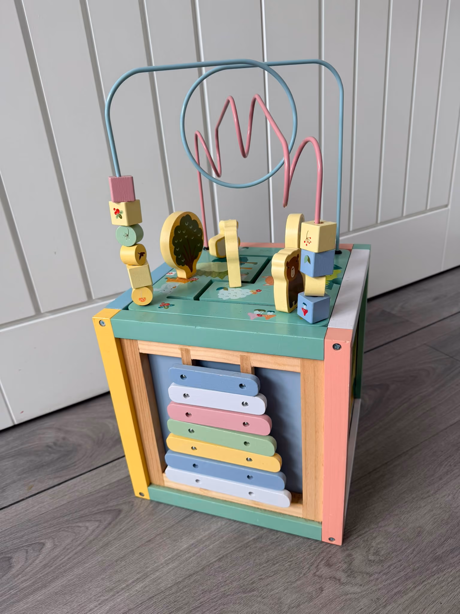 Colorful wooden activity cube with bead maze, shape sorter, xylophone, and animal blocks on a wood floor.
