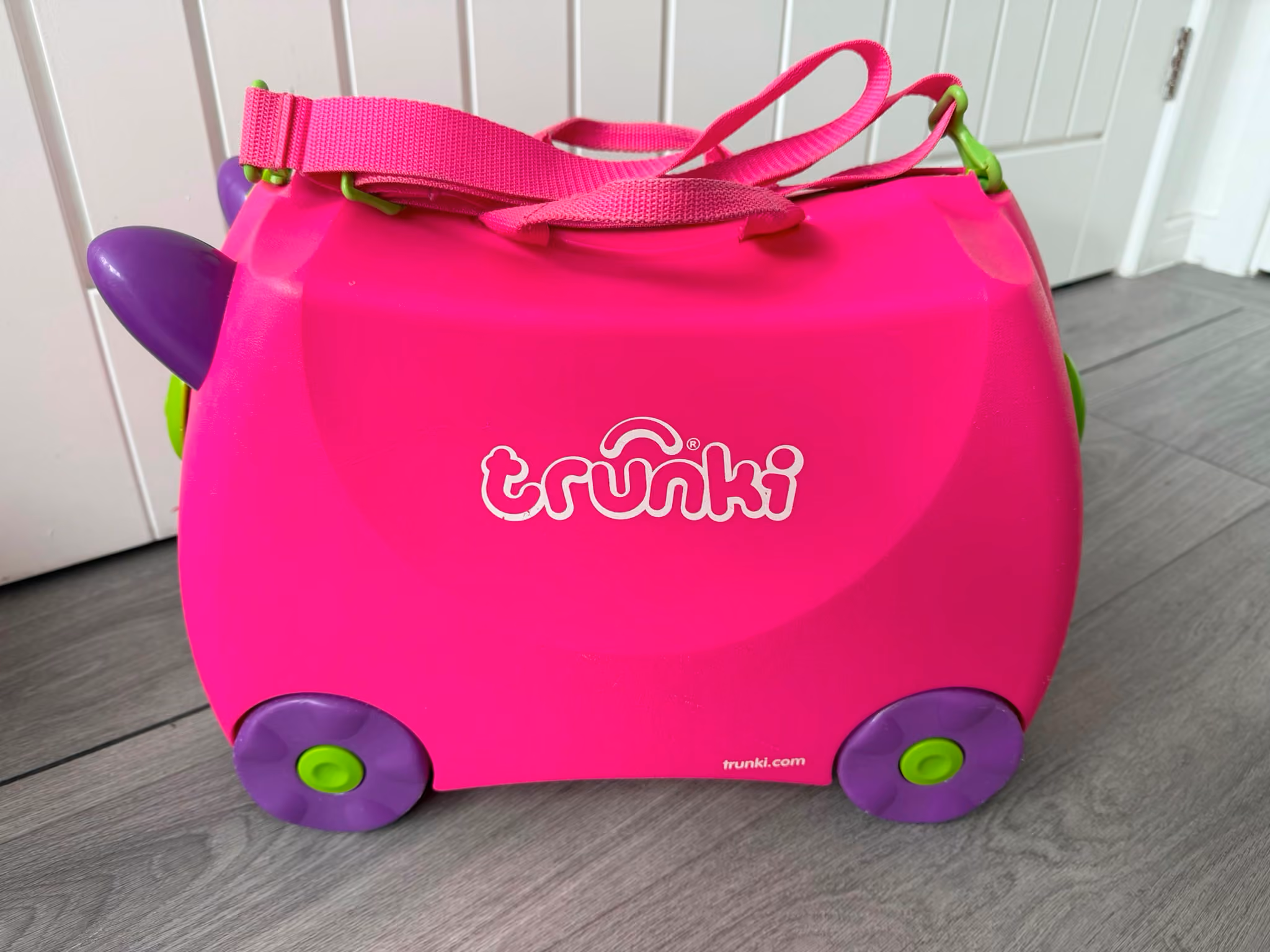 Bright pink Trunki children's ride-on suitcase with purple wheels and a carrying strap indoors.