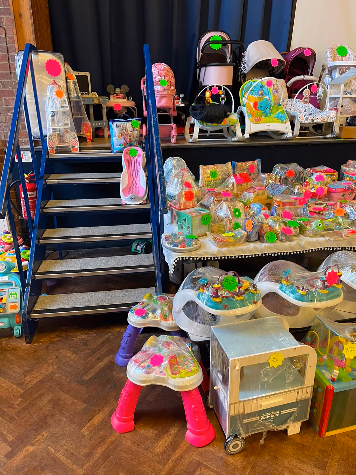 Indoor market stall with baby strollers, toys, and baby gear all marked with colorful price tags.