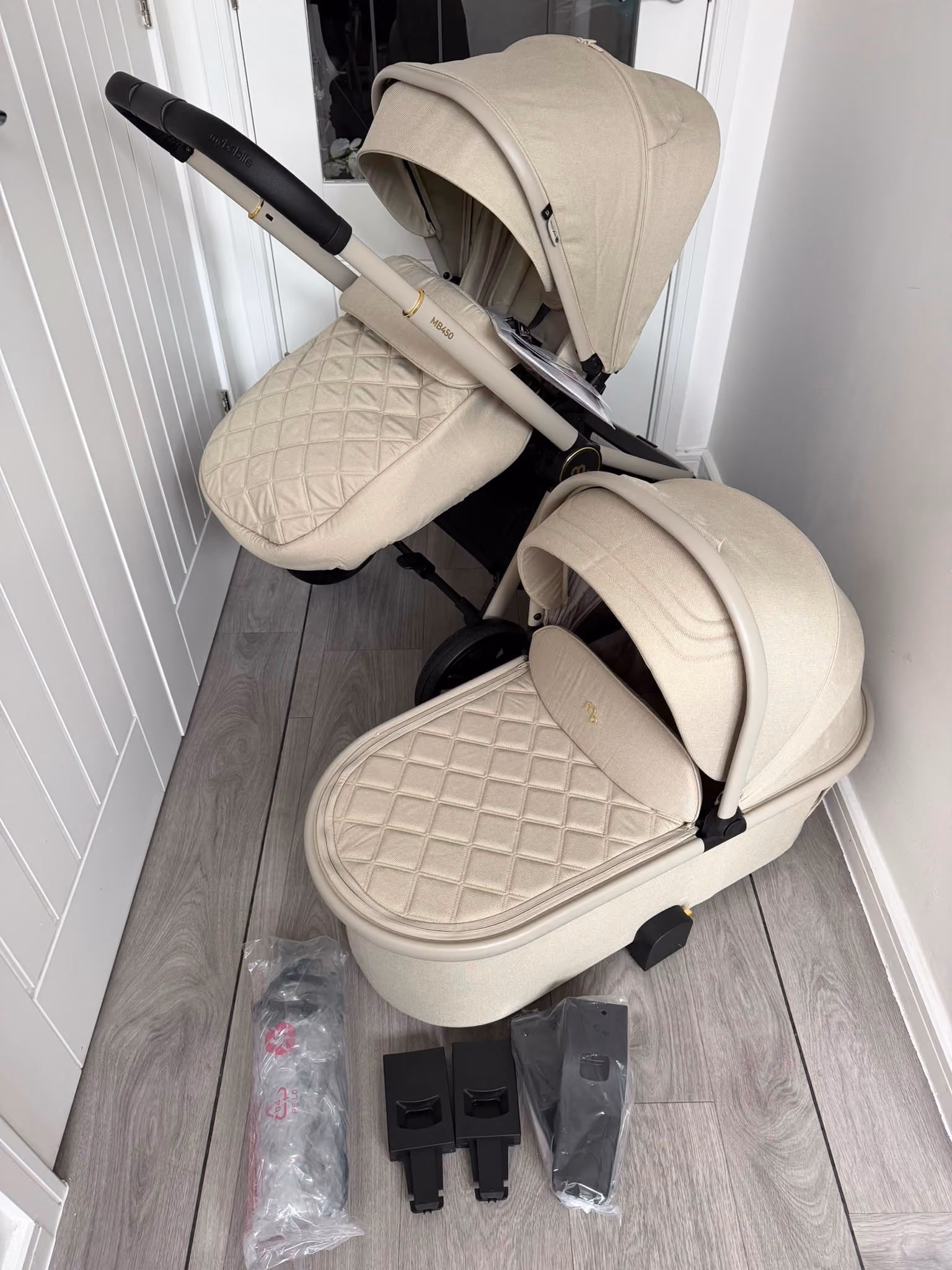 Beige double baby stroller with quilted covers and three black stroller accessory parts on a wood floor.