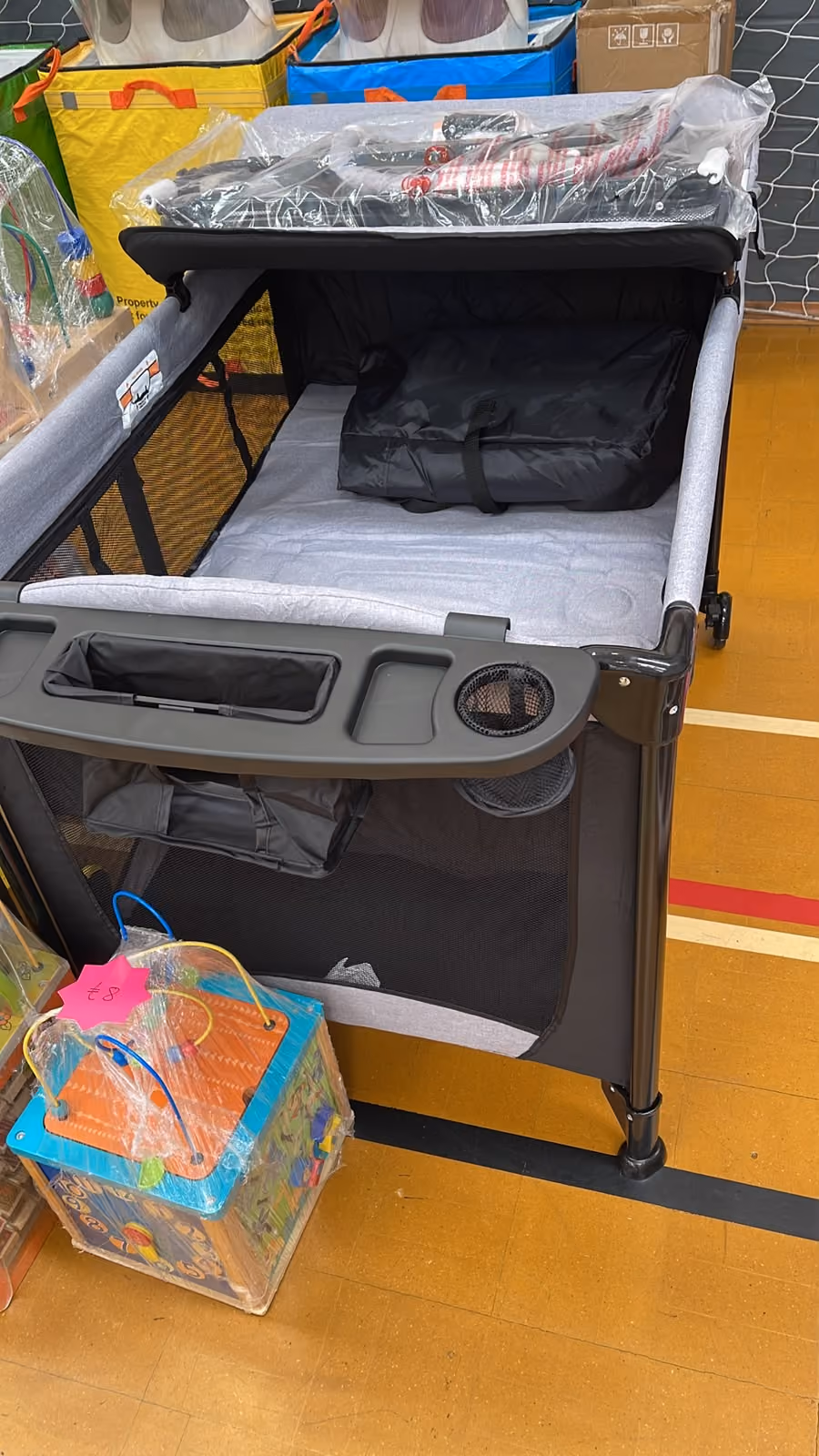 Gray playpen on gym floor with black folded item inside and colorful children's toy wrapped in plastic nearby.