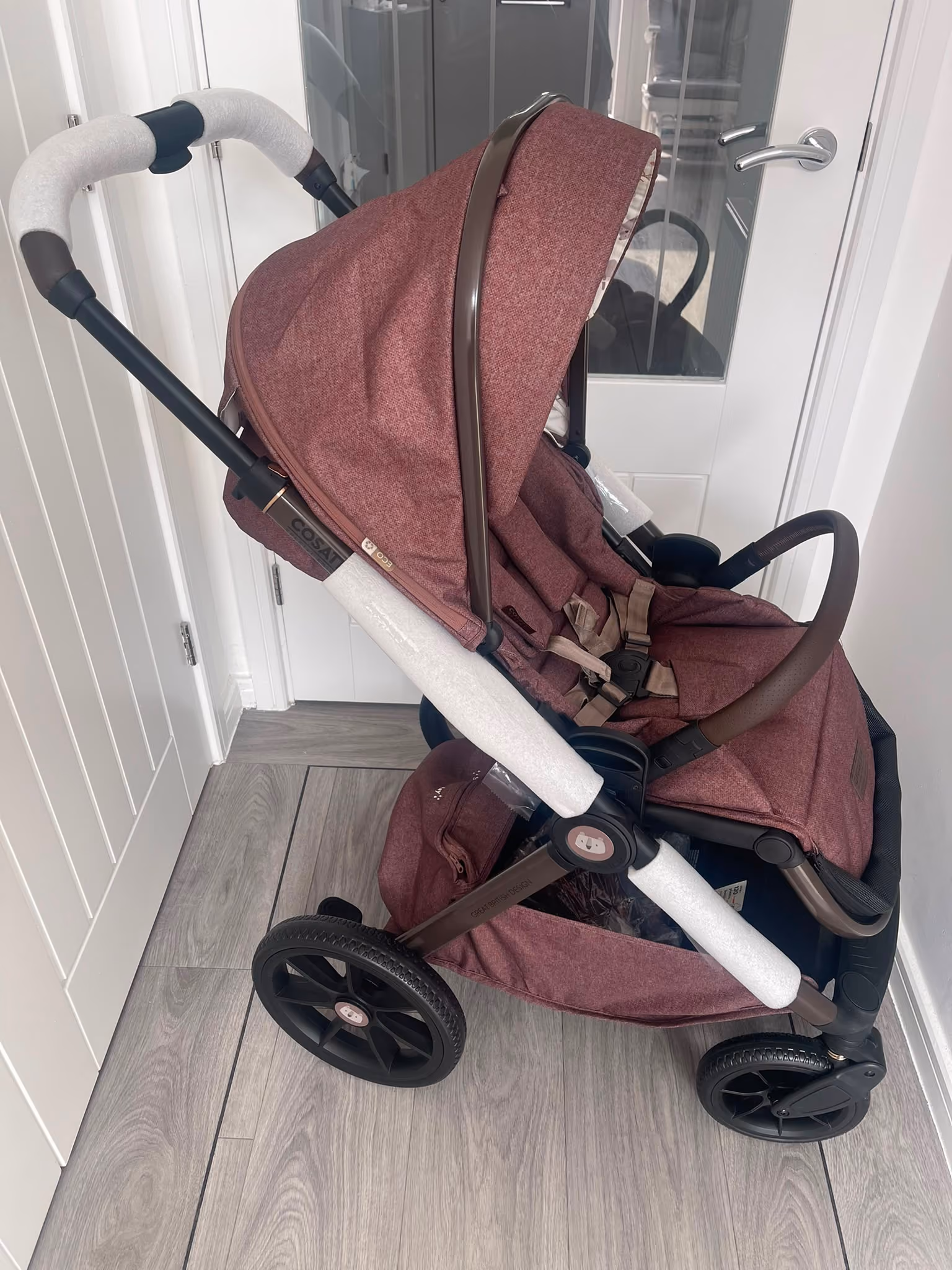 Maroon baby stroller with protective foam on handle and frame, positioned indoors on wood floor.