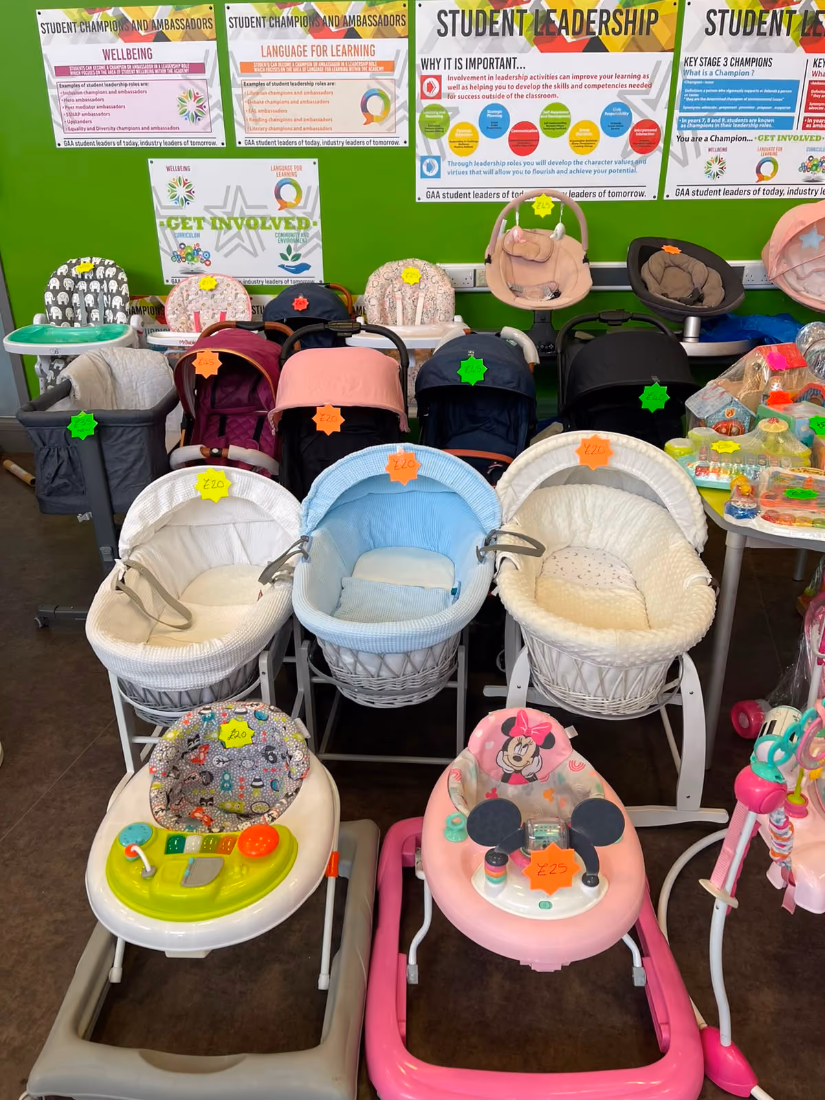 Various baby strollers, carriers, and walkers with price tags displayed indoors against a green wall.