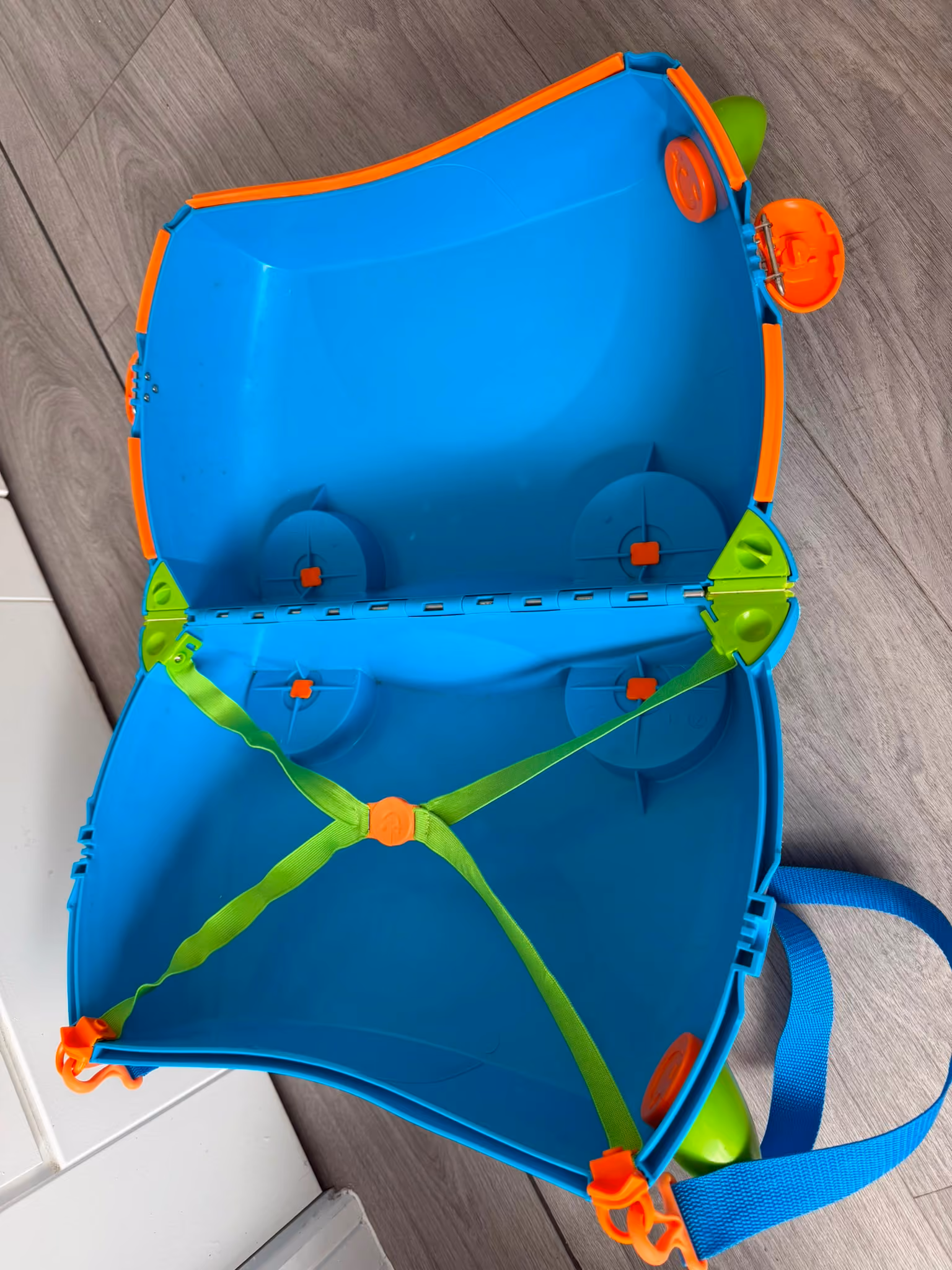 Open blue hard-shell suitcase with orange edges and green straps on wood floor.