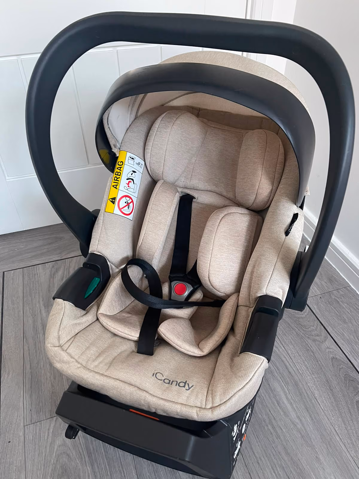 Beige infant car seat with black handle and safety harness on wooden floor.