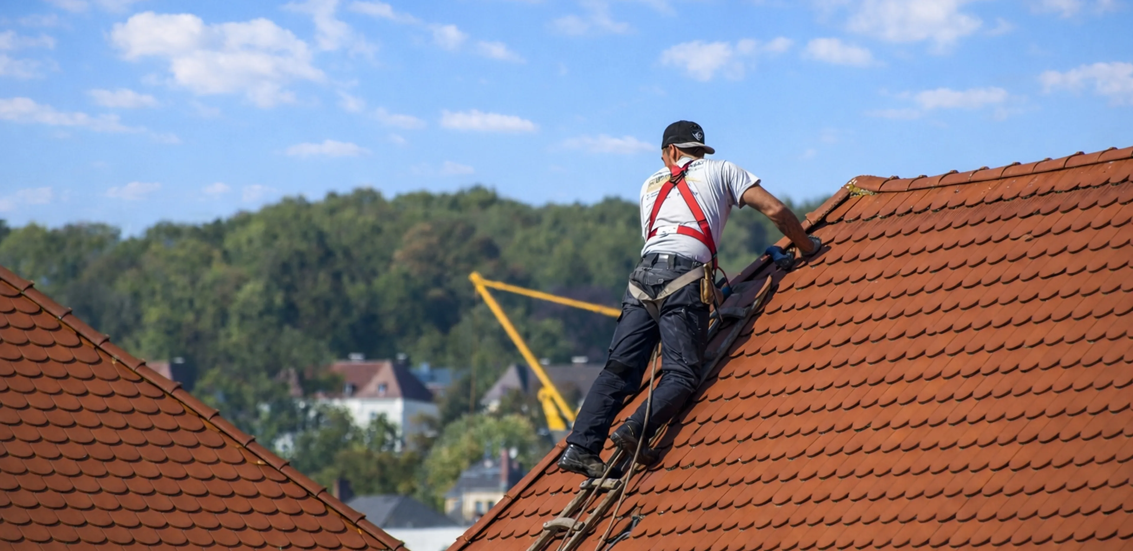 A worker wearing safety harness and cap repairs red tiled roof under a blue sky with trees and houses in the background.
