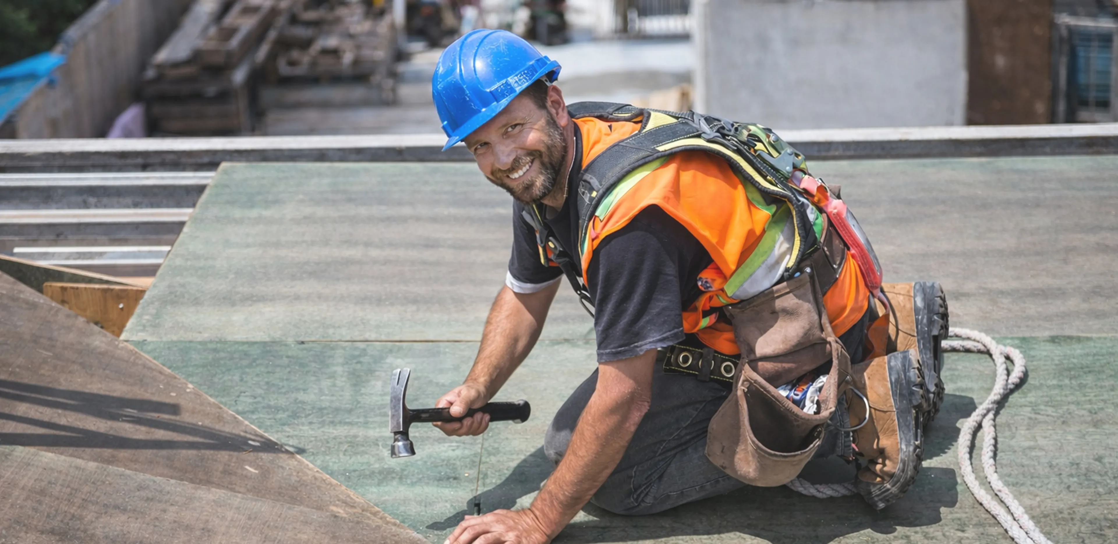 Smiling construction worker wearing a blue hard hat and safety vest kneeling while using a hammer on a building site.