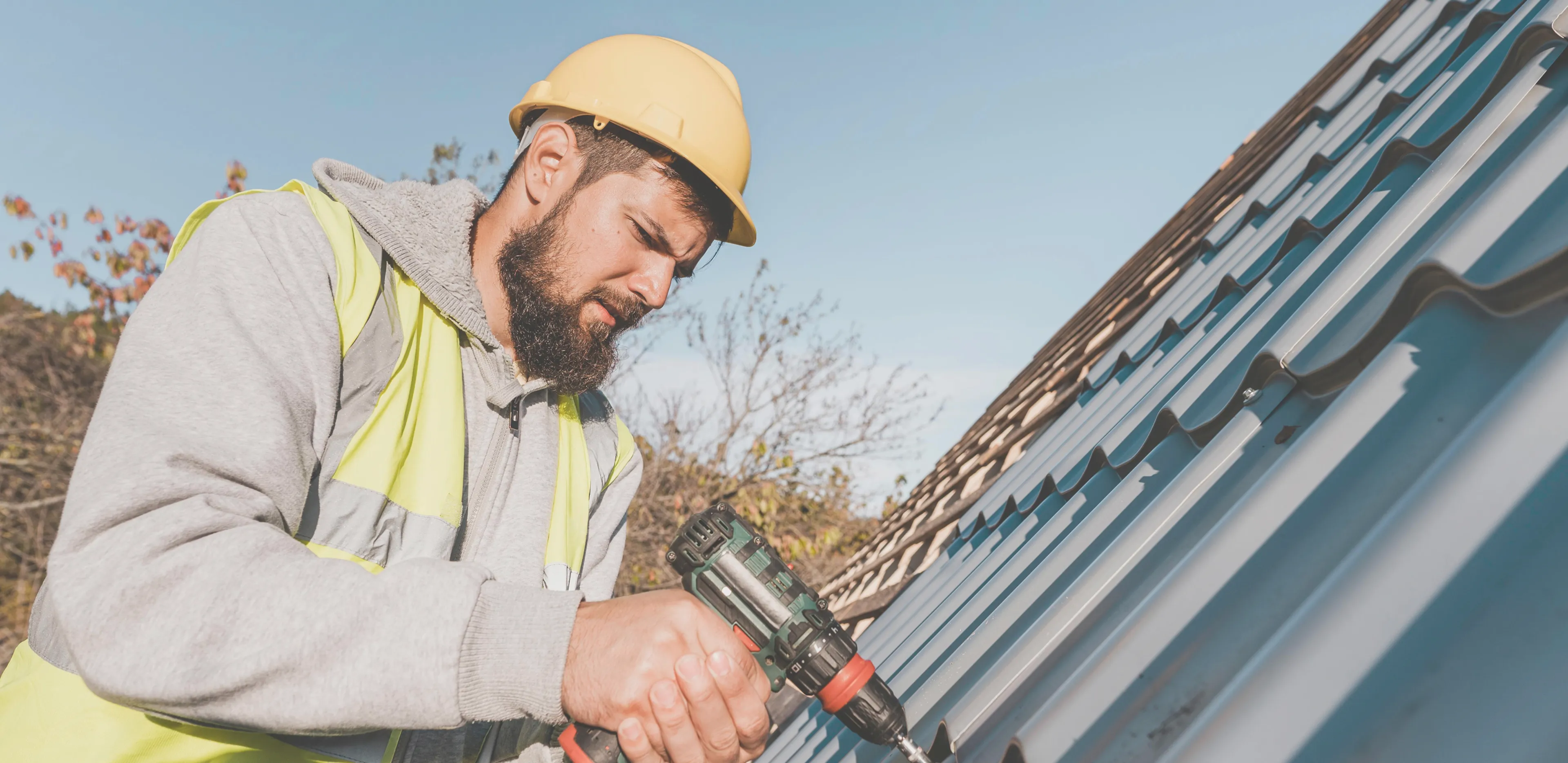 Construction worker wearing a yellow hard hat and safety vest using a power drill on a metal roof.