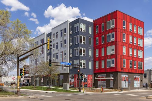Street-corner view of the apartment building clad in red, navy, grey and white panels above ground-floor commercial storefronts
