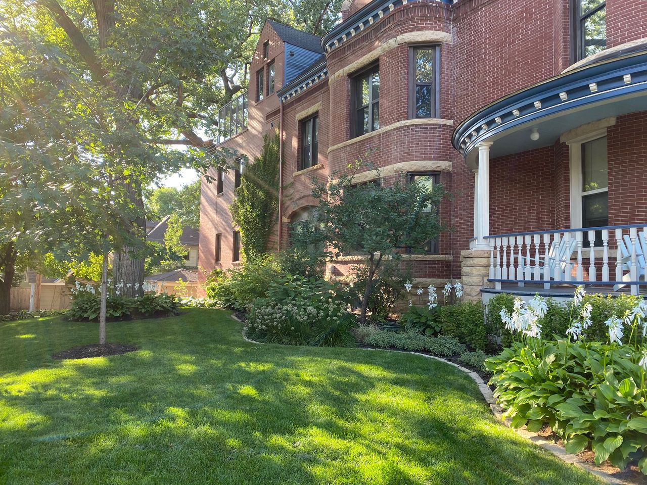 Side lawn with mature trees and planting beds of hostas running alongside the building's brick wall and porch railing