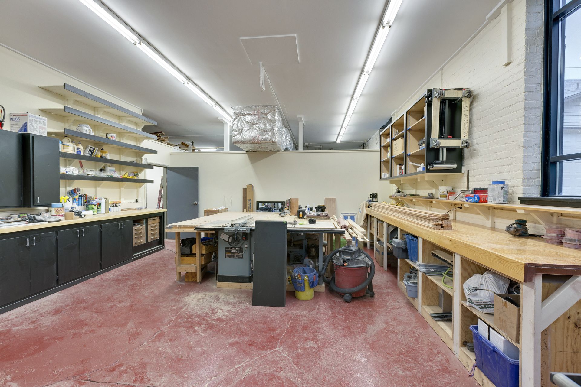 Wide woodworking shop with black base cabinets, open shelving, a table saw and overhead dust collection ducting