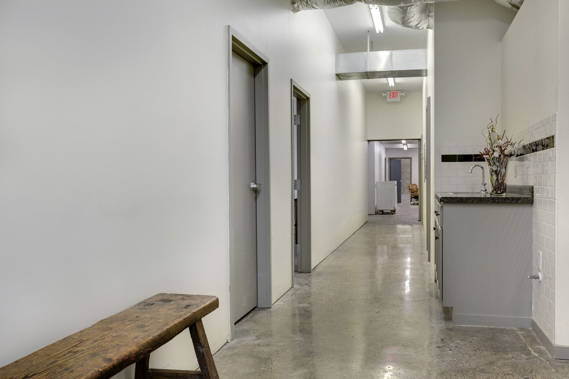 Corridor with grey doors, a long wooden bench, exposed ductwork and a kitchenette visible at the far end
