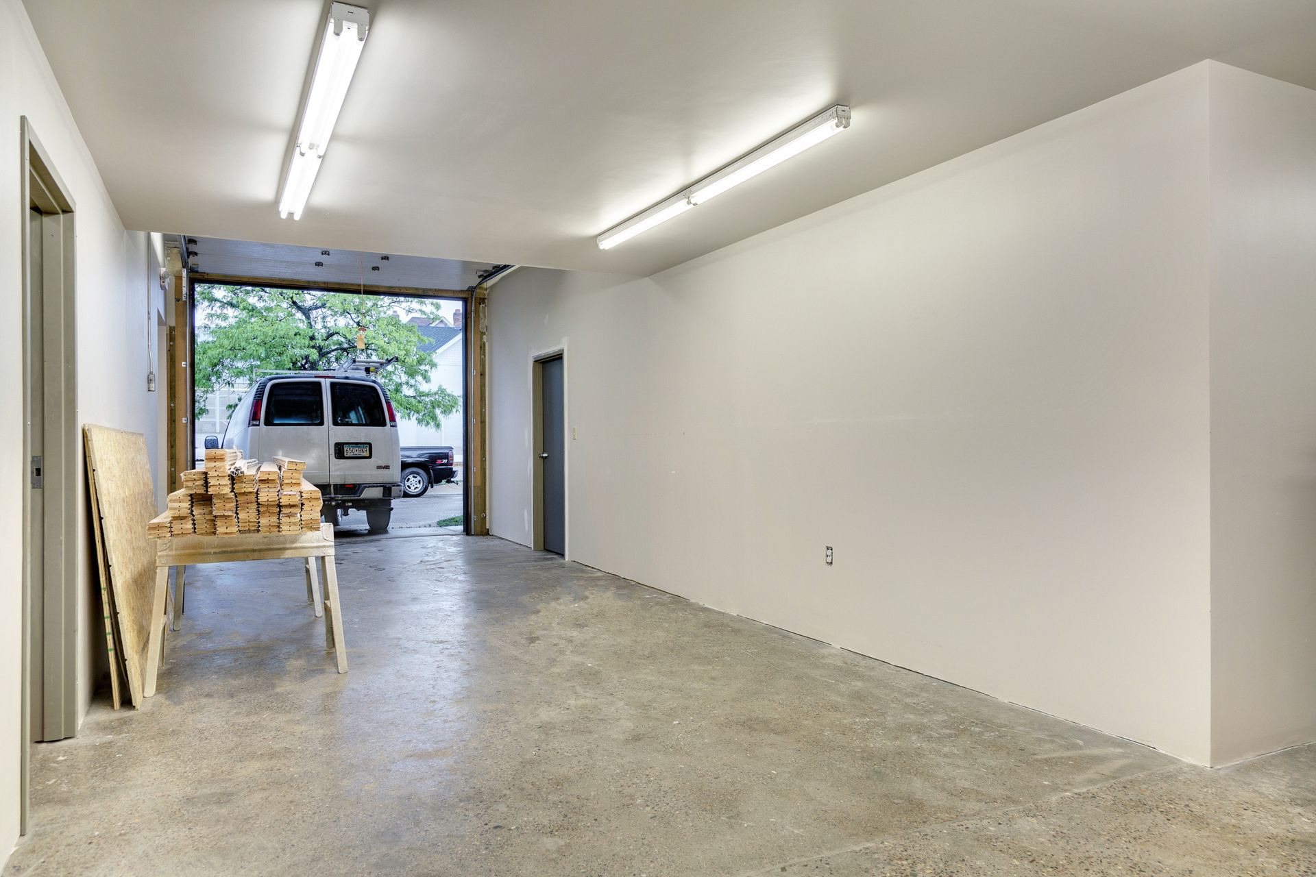 Room with white walls and an open overhead drive-in door, a sawhorse table holding stacked wood blocks and a van parked outside
