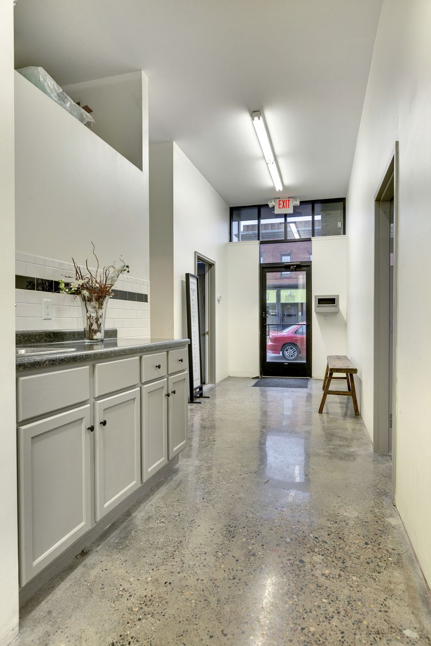 Corridor with a grey-cabinet kitchenette along one wall, polished concrete floor and a glazed entrance door at the far end