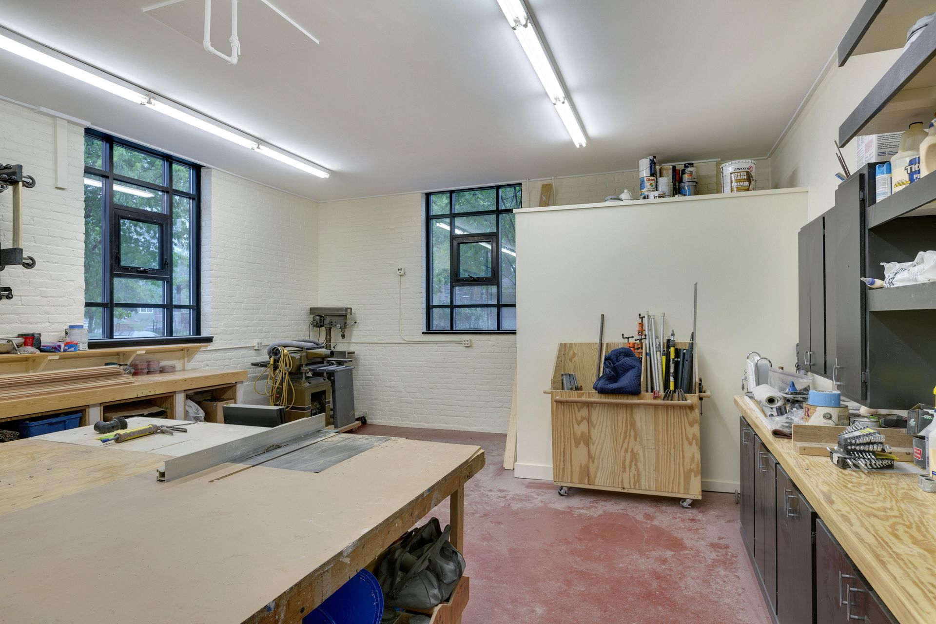 Woodworking shop with white-painted brick walls, black-framed windows, a table saw and a mobile lumber cart on a red sealed floor