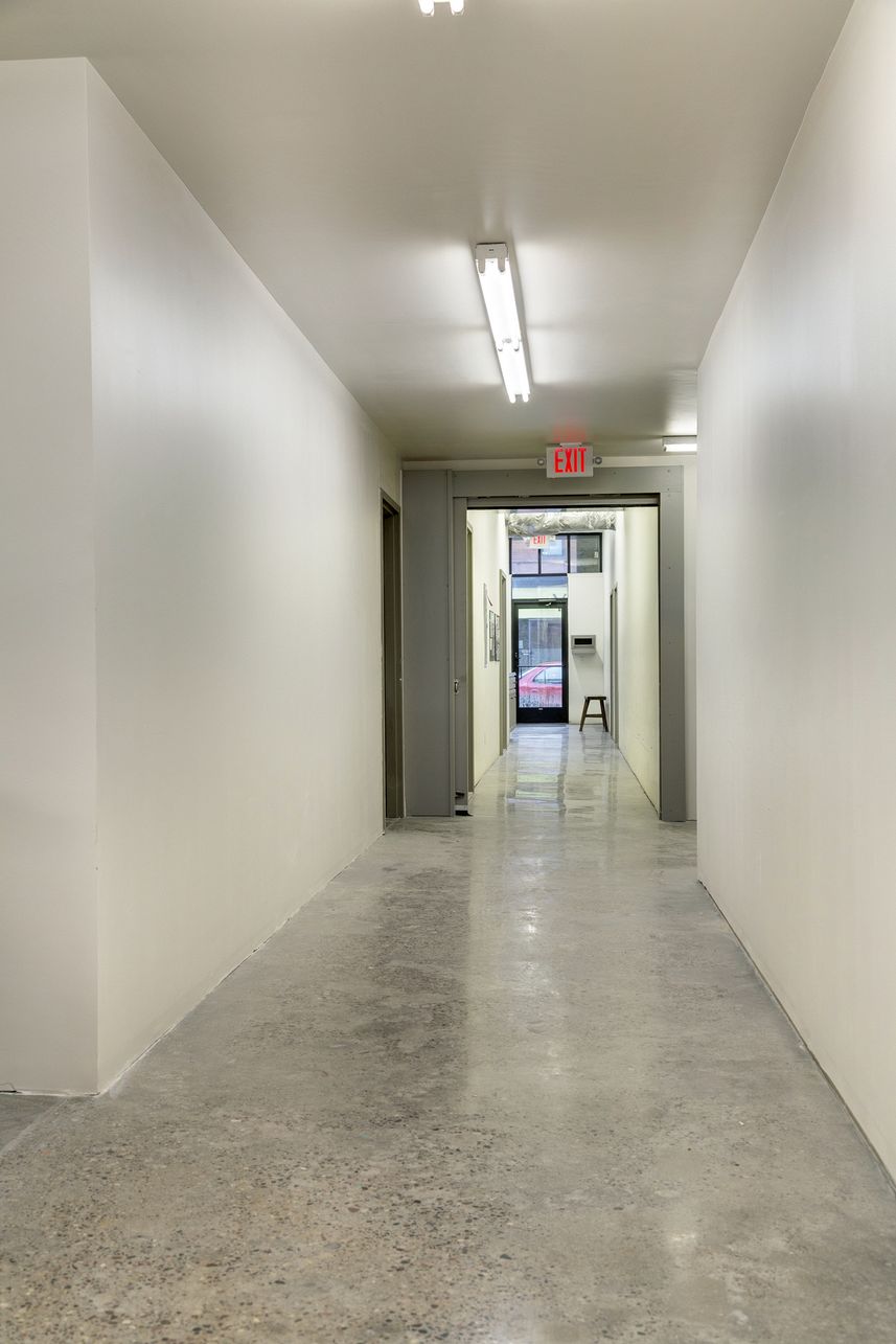 Corridor with white walls and a polished concrete floor leading to a glazed entrance door beneath an exit sign