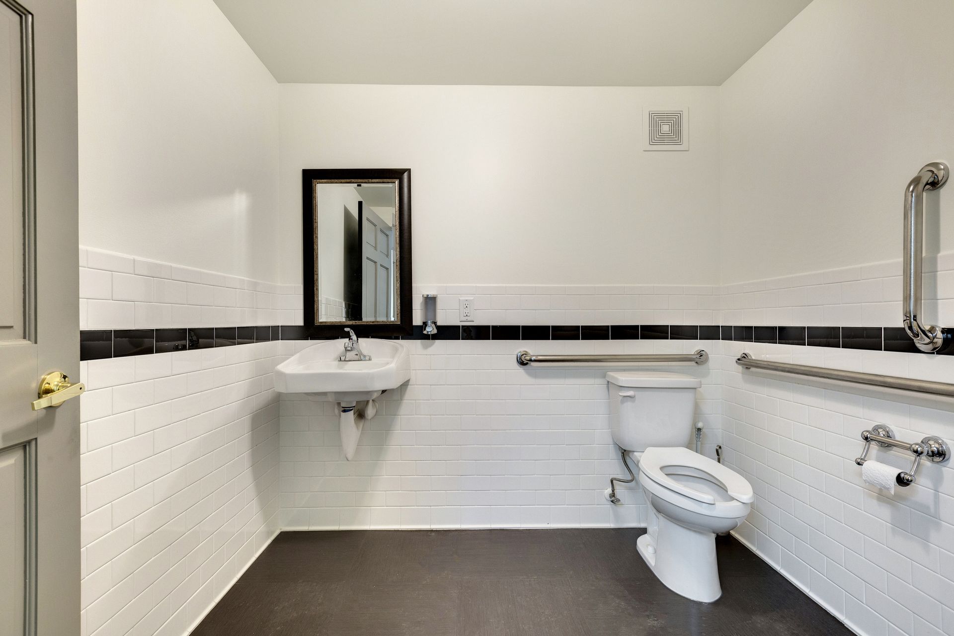 Restroom with white subway tile, a black accent band, a wall-mounted sink, a framed mirror and grab bars beside the toilet