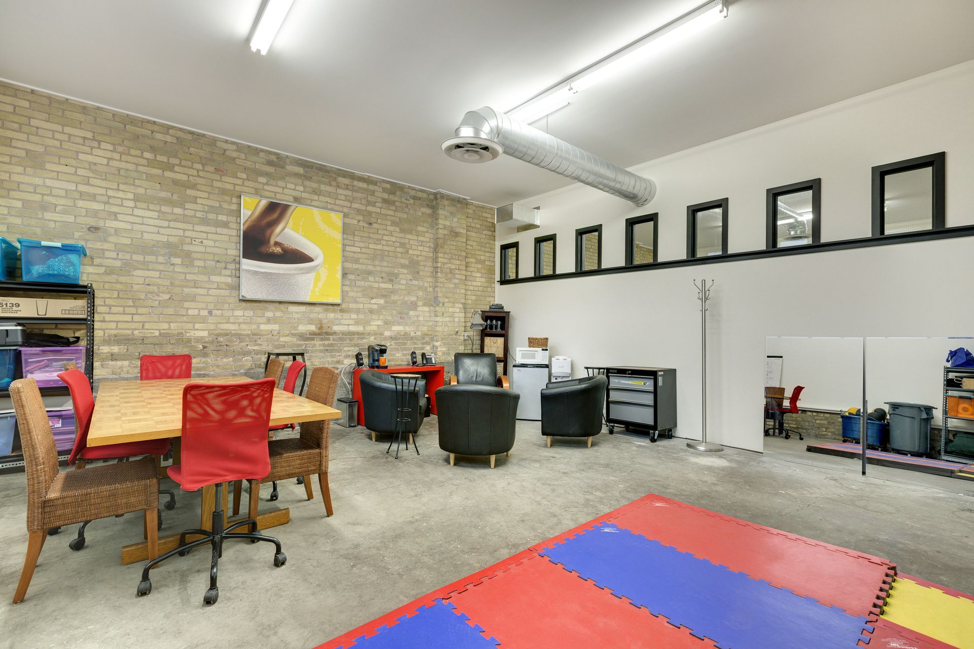 Shared room with clerestory windows, storage shelving, a flip chart, a table with red chairs and exposed spiral ductwork