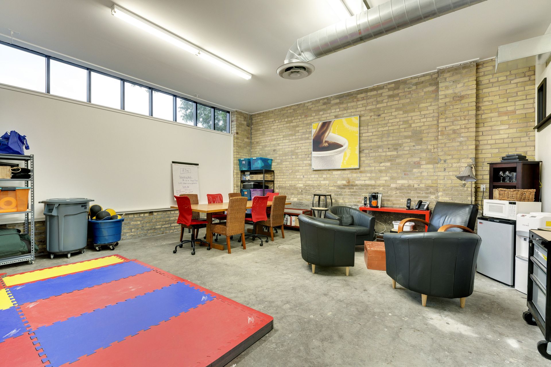 Shared room with a wooden table and red chairs, black tub armchairs, framed artwork and interlocking foam floor mats