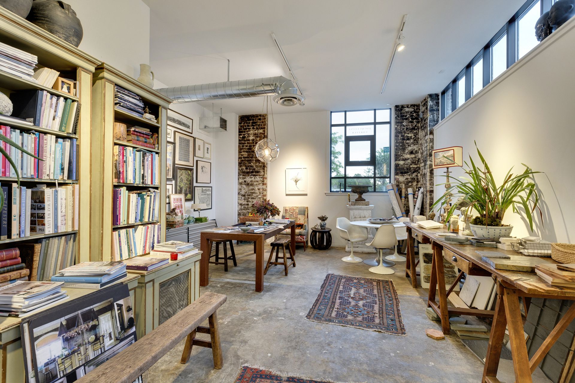 Studio with tall painted bookcases full of books, trestle work tables, an exposed brick pier and patterned rugs on a concrete floor
