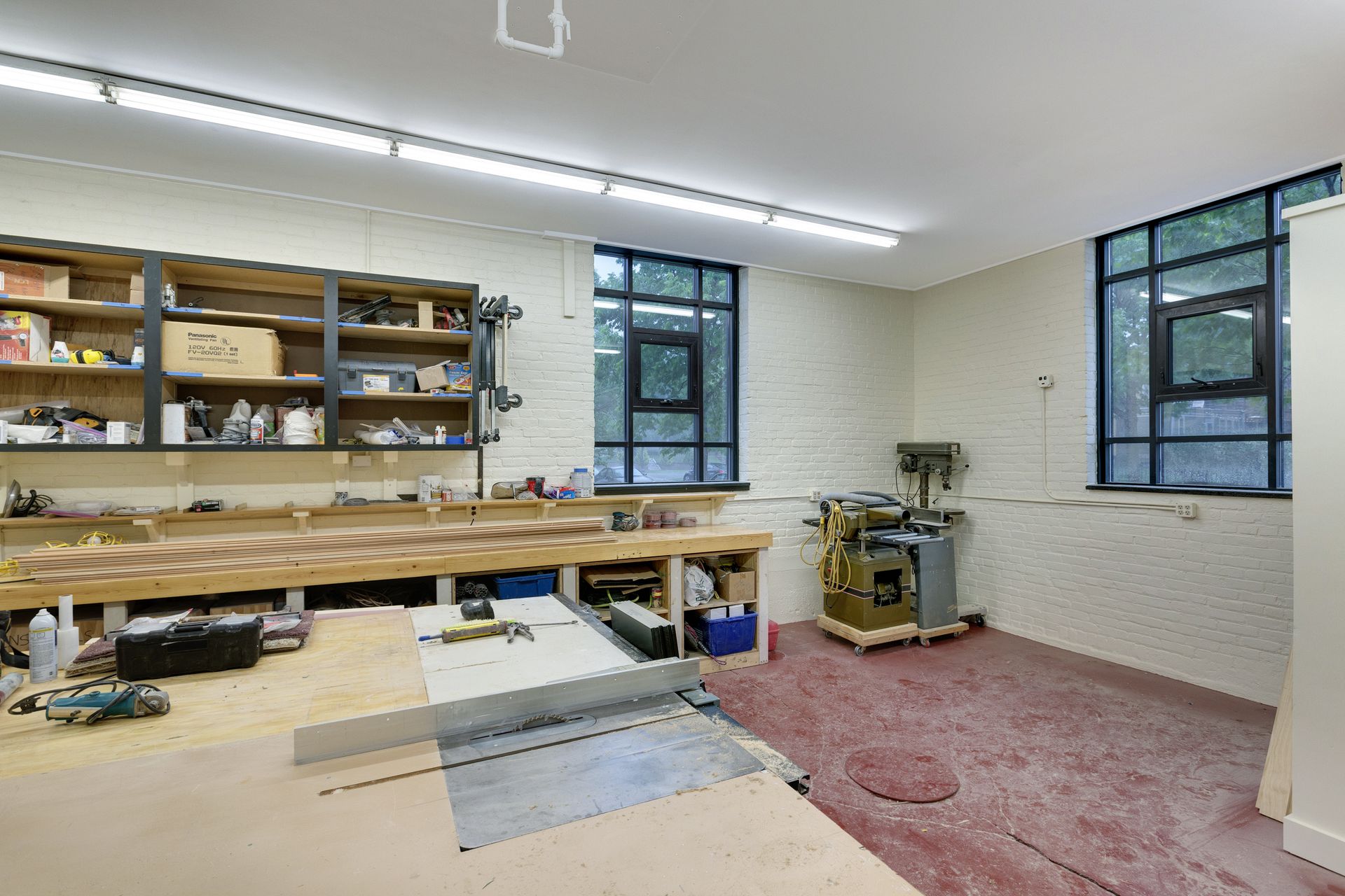 Woodworking shop with a long bench, open shelving, a drill press and a table saw on a red sealed floor