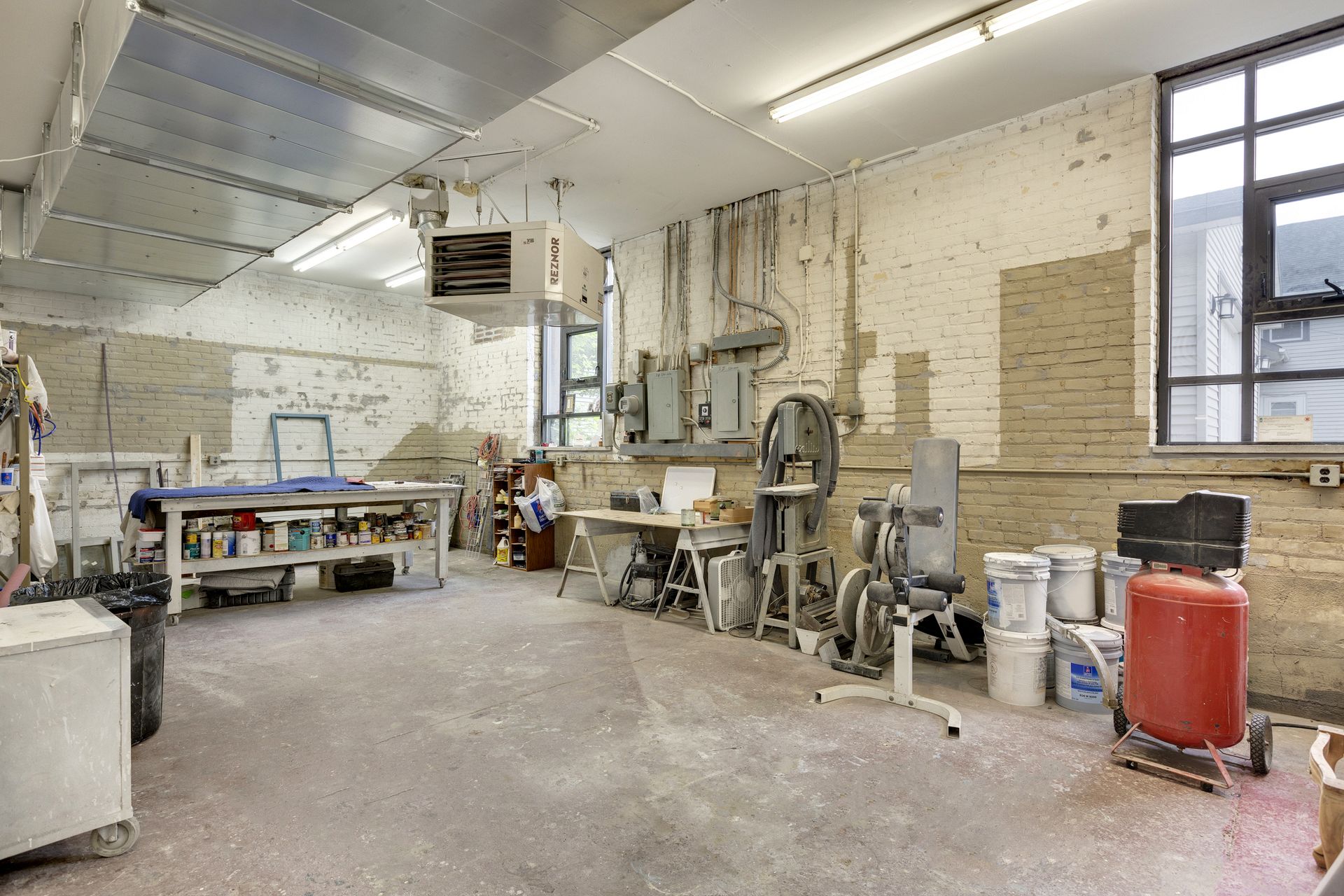 Workshop with painted brick walls, a bandsaw, a red compressor tank, stacked buckets and an overhead gas unit heater
