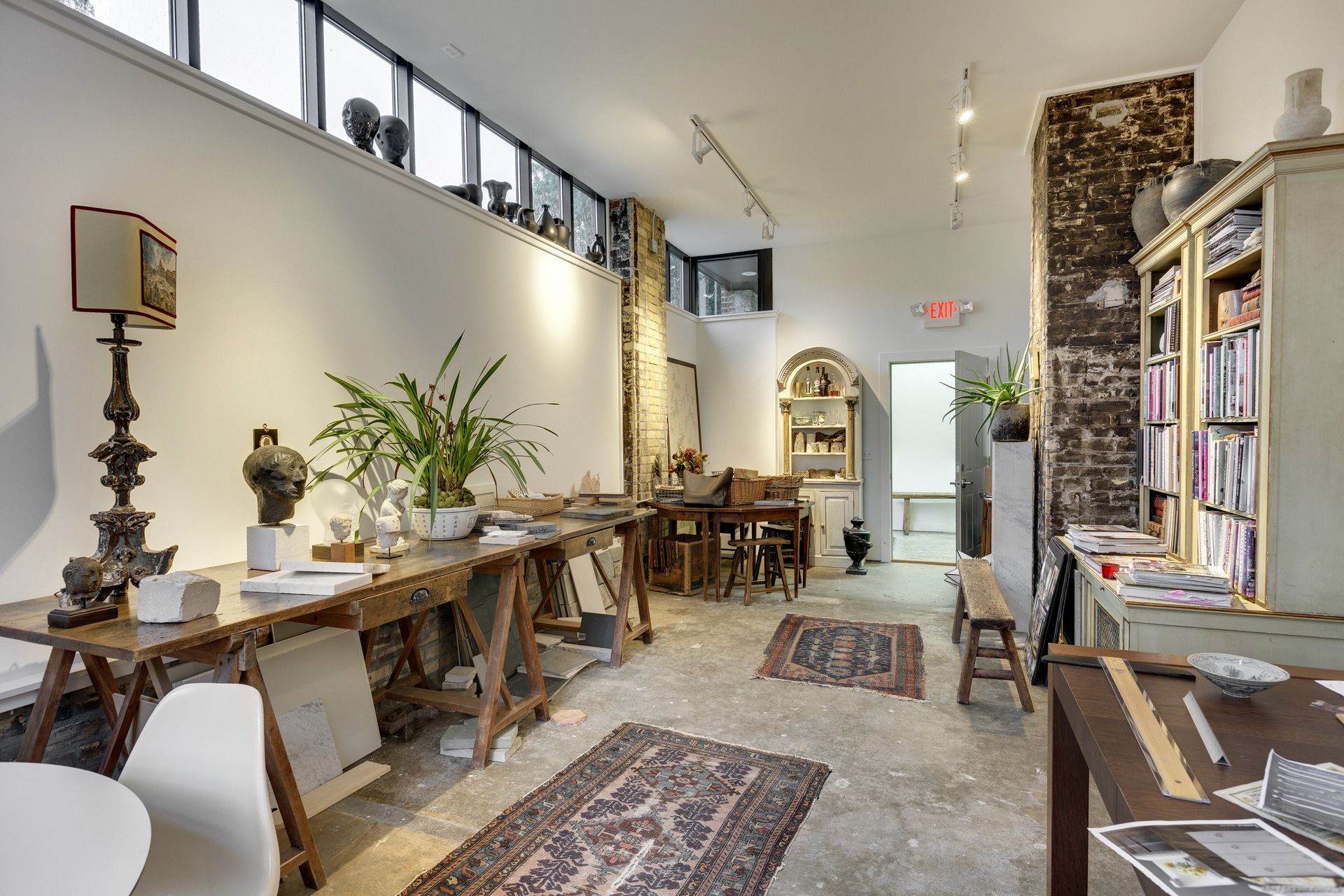 Long studio room with trestle tables, potted plants, framed artwork, clerestory windows and an exposed brick chimney breast