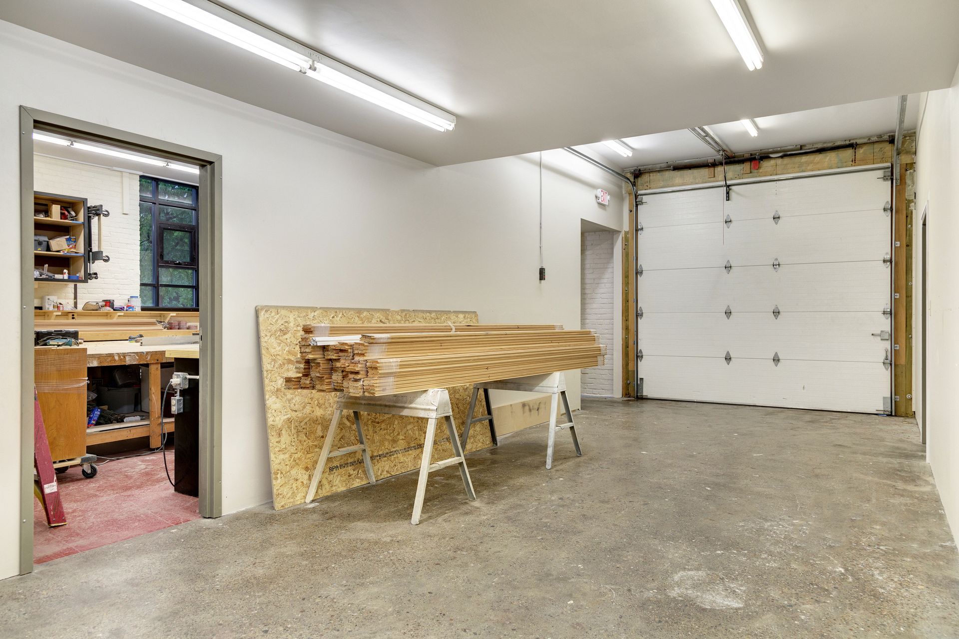 Room with a closed white overhead drive-in door, lumber on sawhorses and a workshop visible through an adjoining doorway
