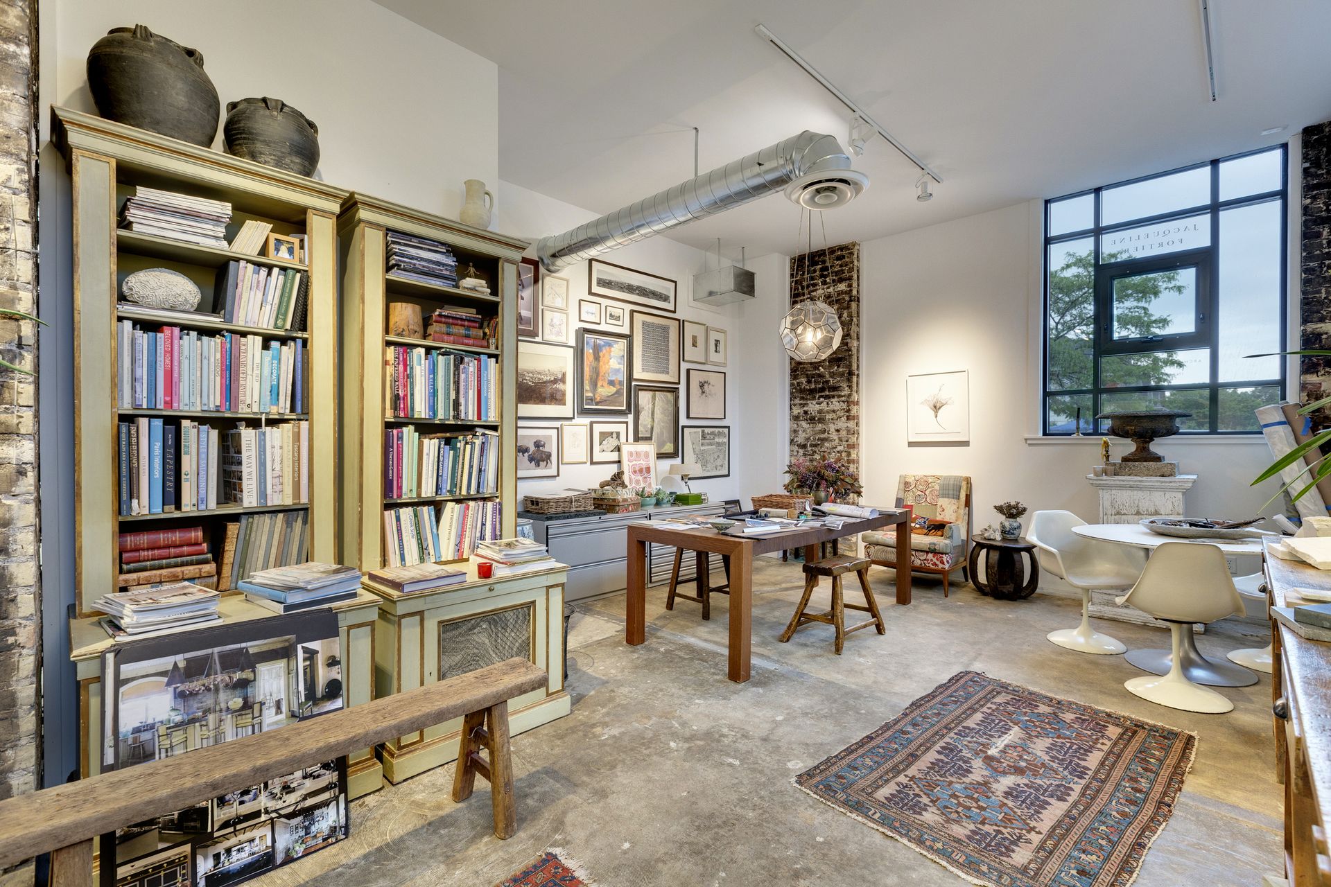 Studio with loaded bookcases, a large black-framed window, a wooden worktable and white tulip chairs