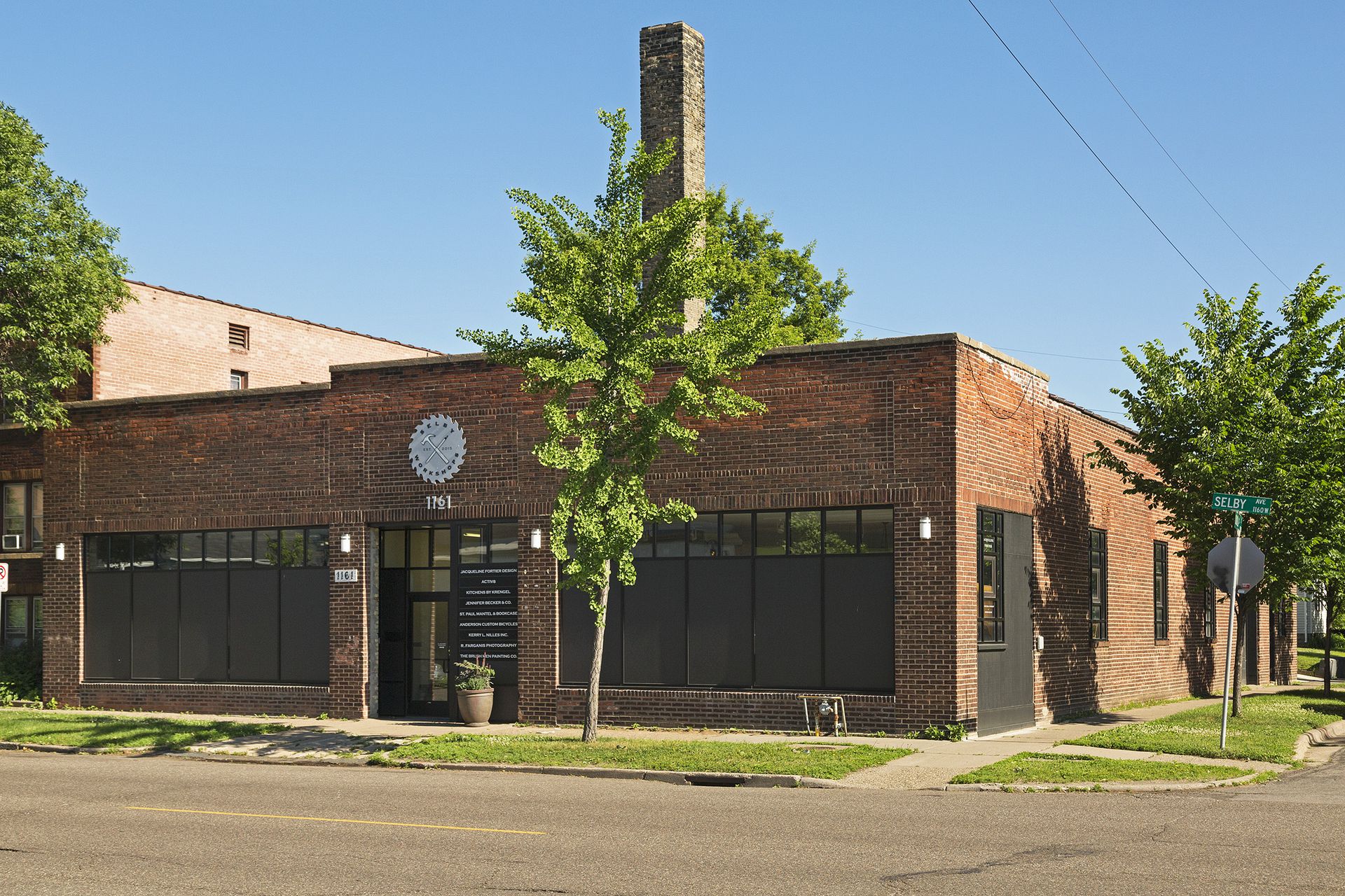 Corner view of a single-storey red brick building with black storefront windows and a tall brick smokestack, seen from across the street
