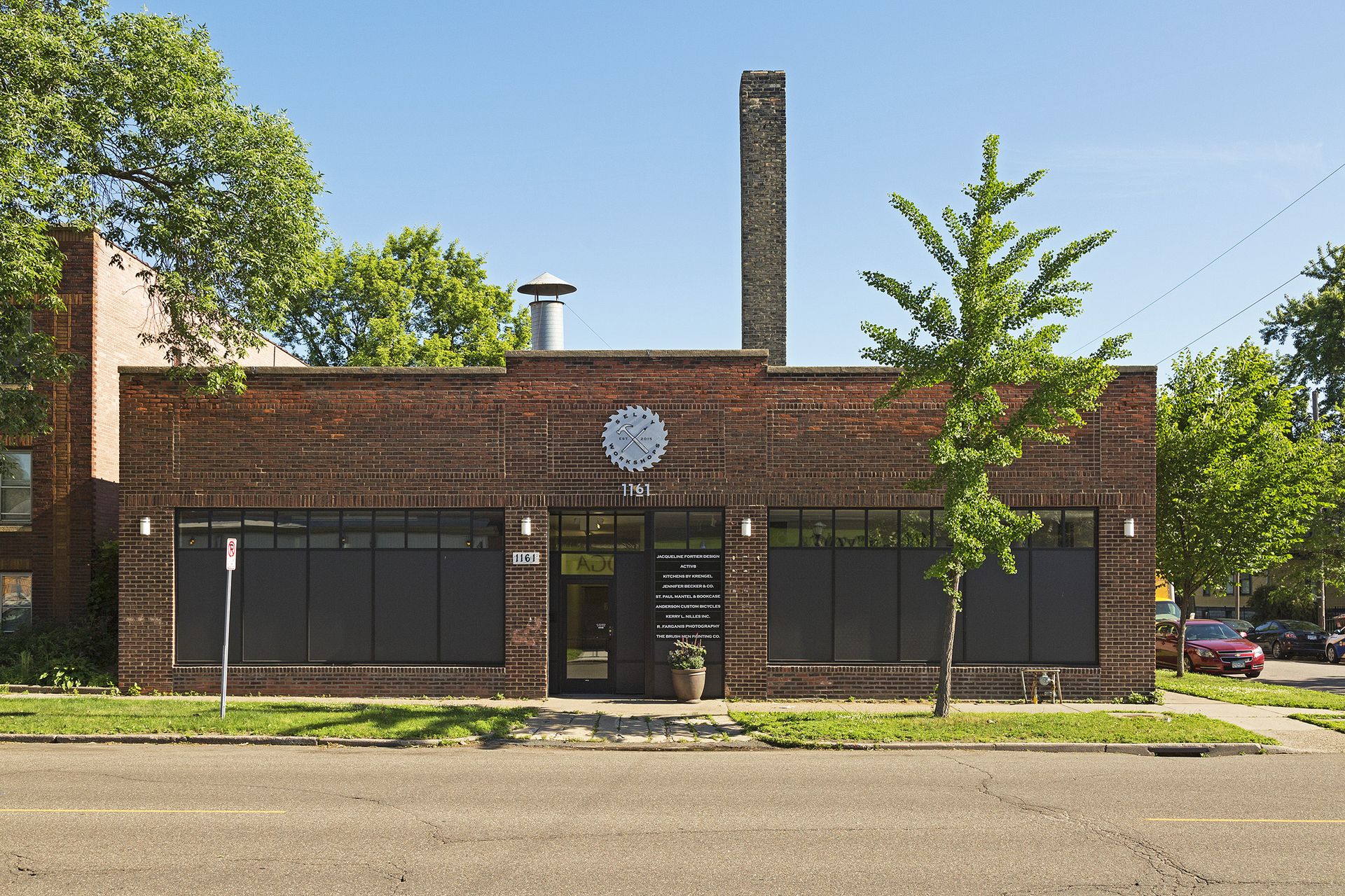 Front elevation of the Selby Workshops Building, a low red brick building with black-framed window bands and a tall brick smokestack
