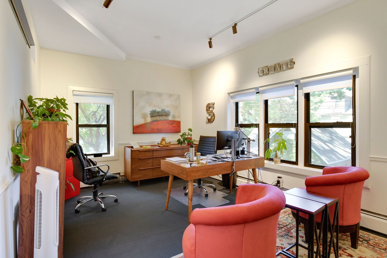 Private office with a wooden desk, coral upholstered tub chairs, framed artwork and tall windows with dark wood trim
