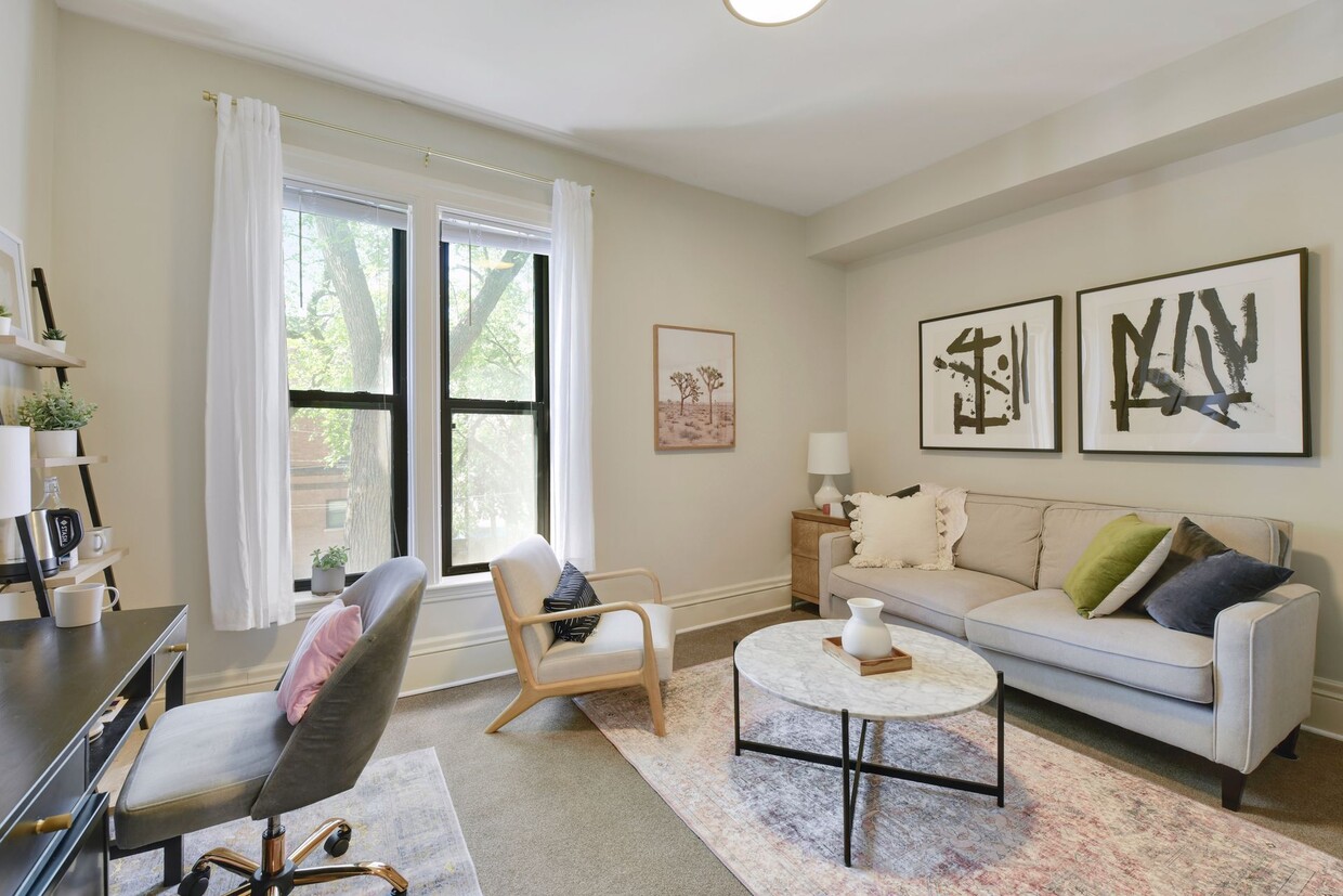 Staged office suite with a grey sofa, a tall black-framed double-hung window with white curtains, framed prints and a desk
