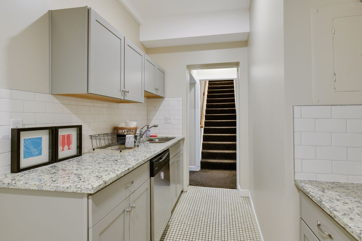 Galley kitchenette with grey cabinets, granite countertops, white subway tile and a dishwasher, opening onto a carpeted staircase
