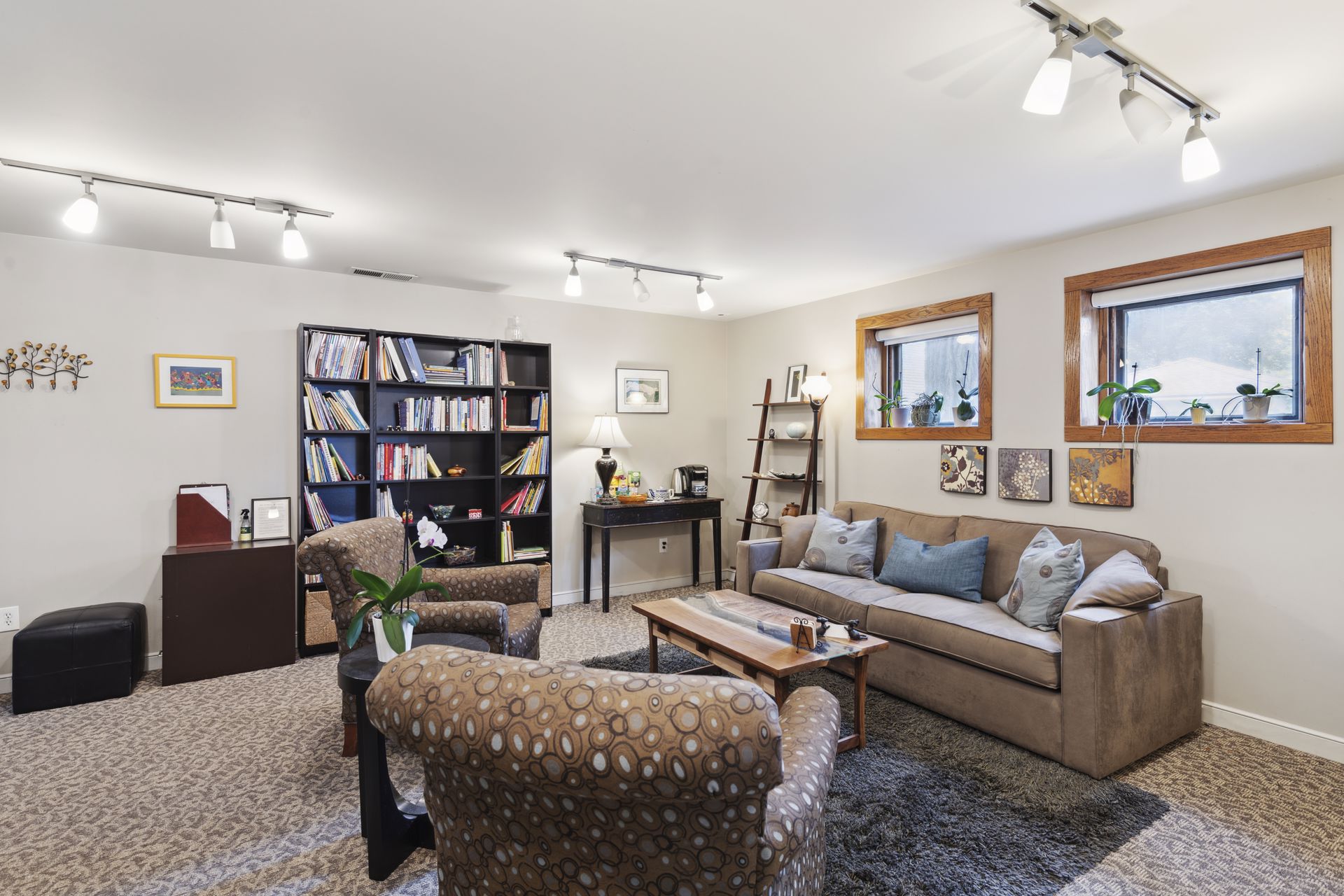 Garden-level lounge with tall black bookcases, a sofa and patterned armchairs beneath wood-framed windows