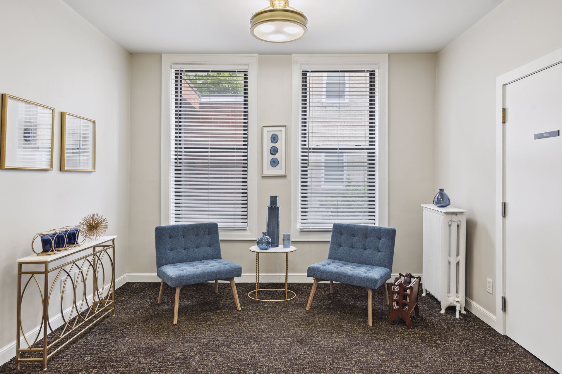 Seating nook with two teal tufted chairs flanking a small round table below two blinded windows