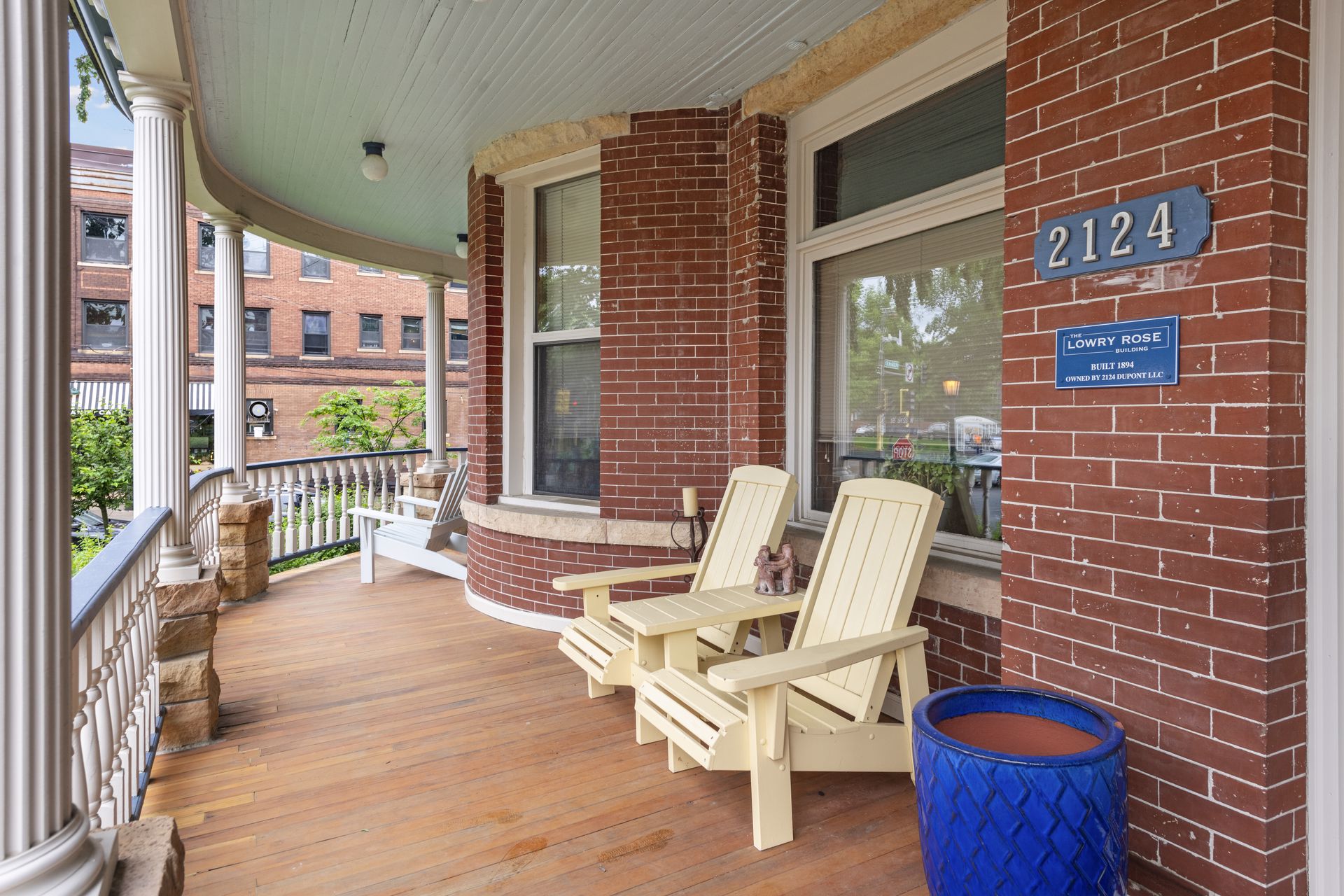 Covered front porch with white columns, a painted wood deck, wooden Adirondack chairs and a blue 2124 address plaque on a brick pier