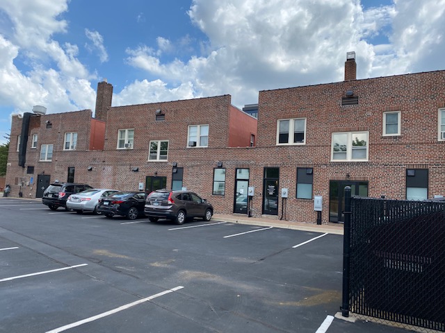 Rear elevation of the brick building seen across a striped surface parking lot with cars parked along the wall