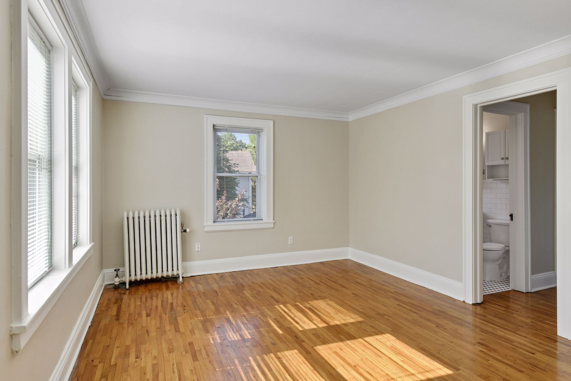 Empty room with a single window and a cast-iron radiator, with a doorway into a tiled bathroom