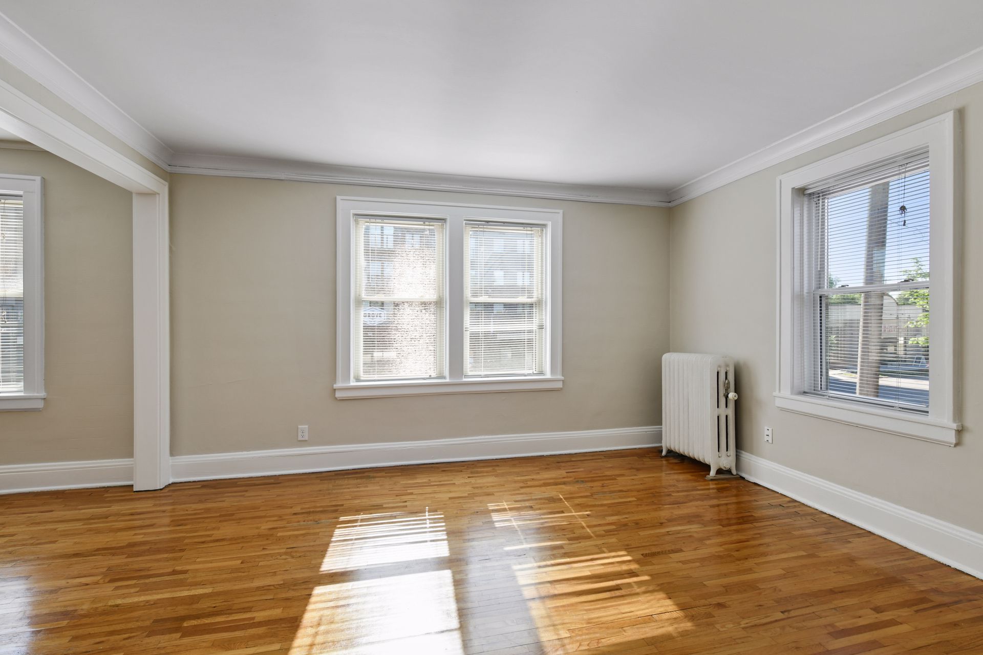 Empty corner room with windows on two walls, a cast-iron radiator and a sunlit oak floor