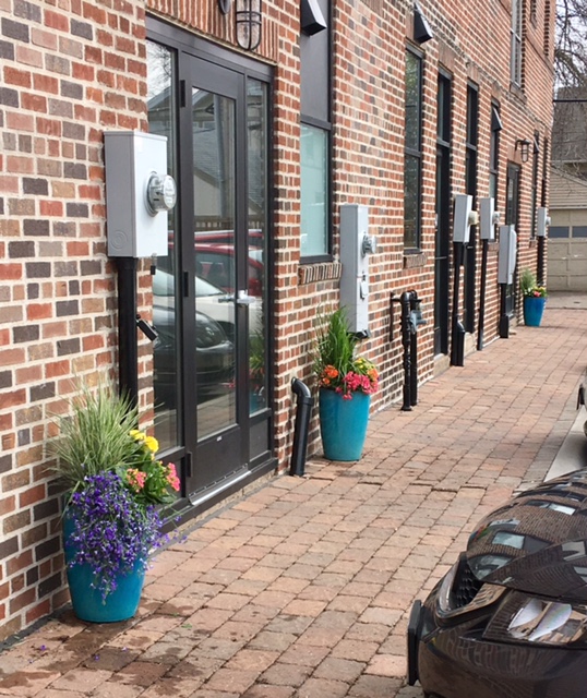 Brick paver walkway beside the building with a glass entry door, wall-mounted utility meters and planters of flowers