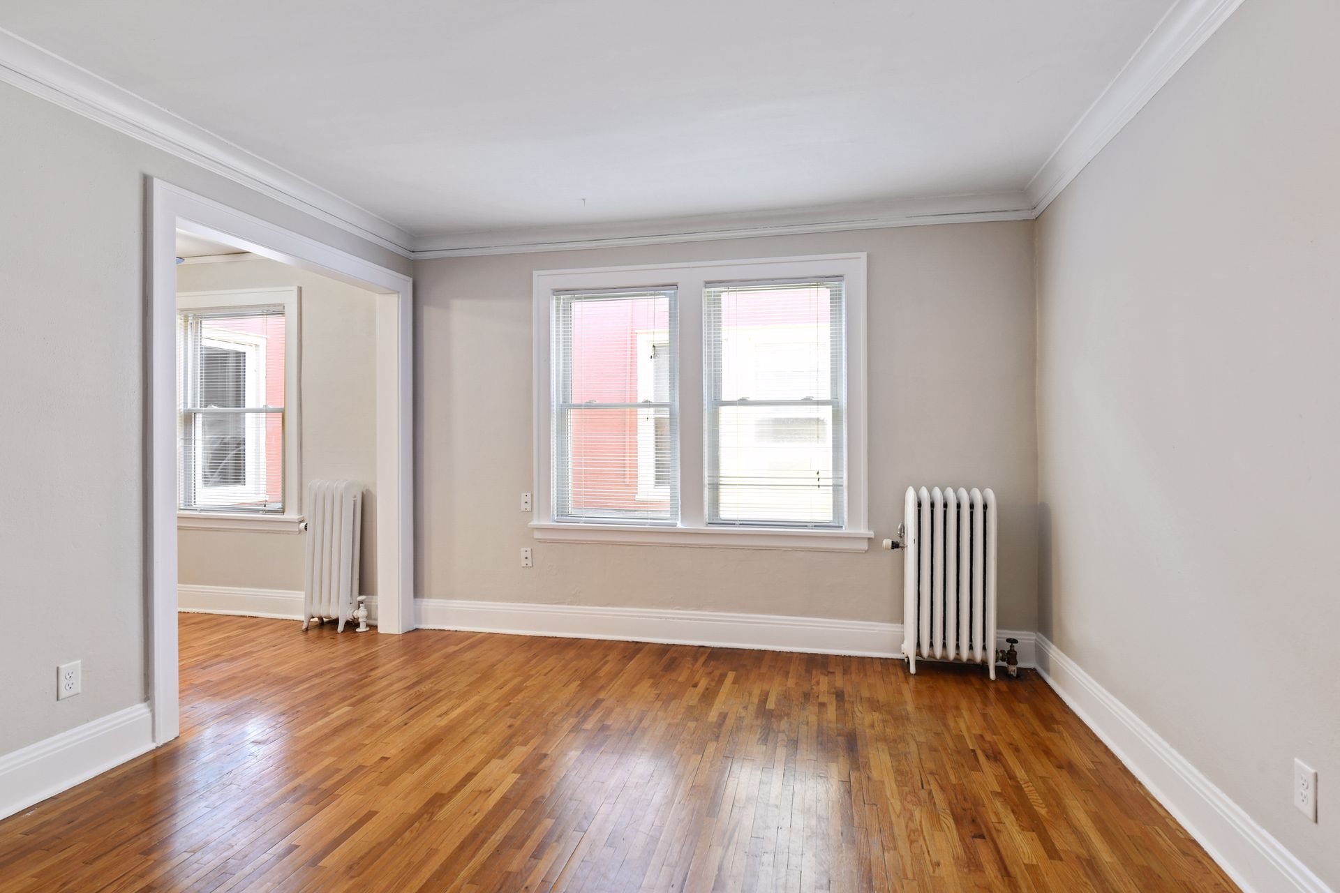 Empty living room with an oak floor, two double-hung windows, two cast-iron radiators and an opening to an adjoining room