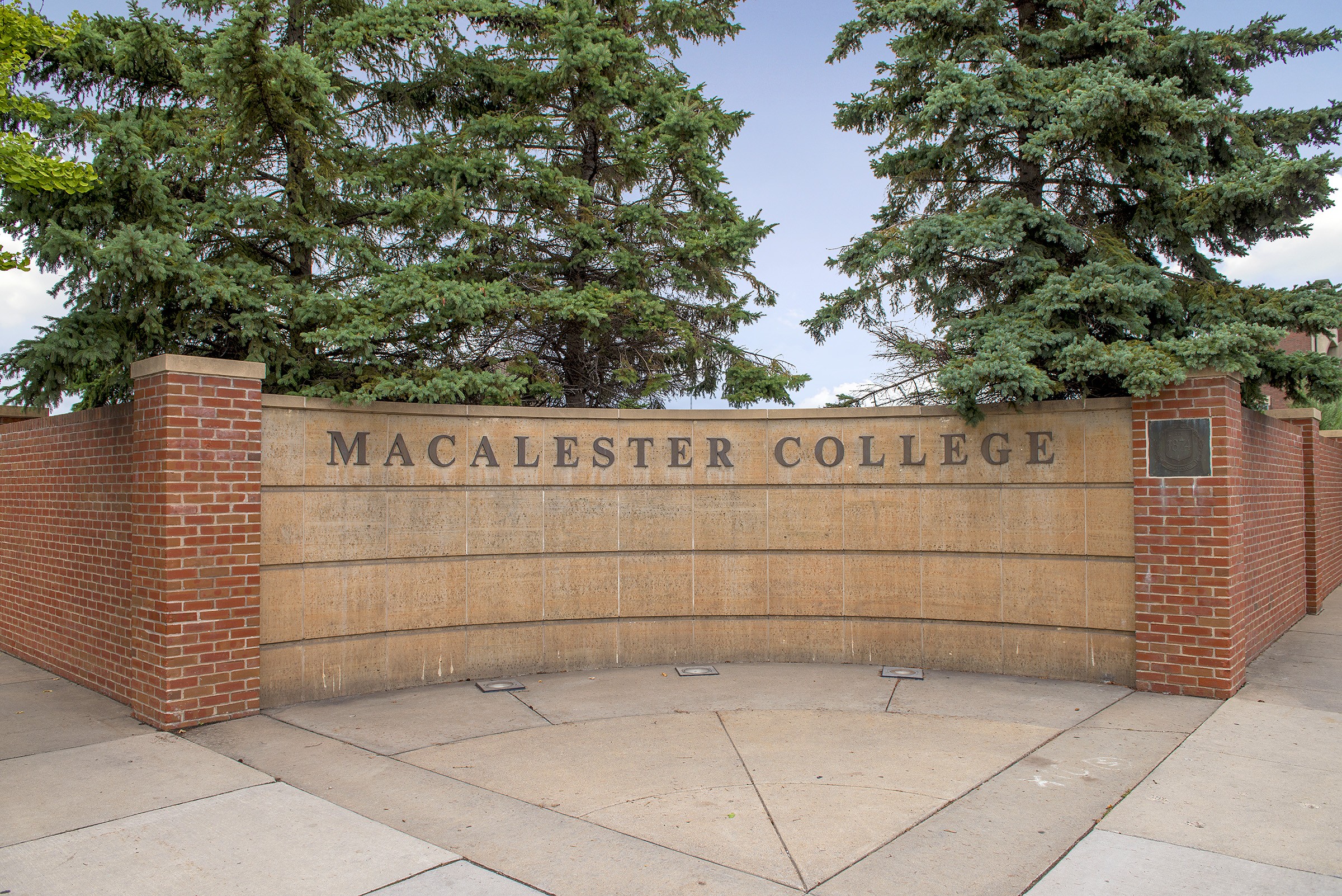 Brick and stone entrance wall lettered Macalester College with evergreen trees behind it