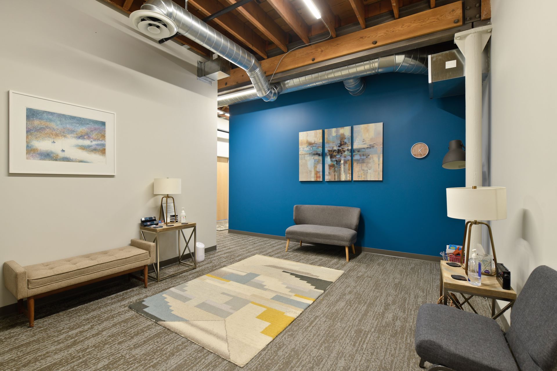 Seating area with a blue accent wall, framed artwork, a bench and sofa, exposed wood joists and a spiral duct overhead