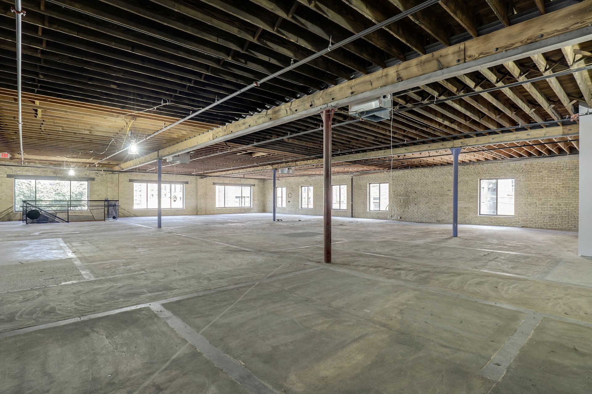 Empty floor plate with exposed wood joists, a brick pier and a stair rail at left