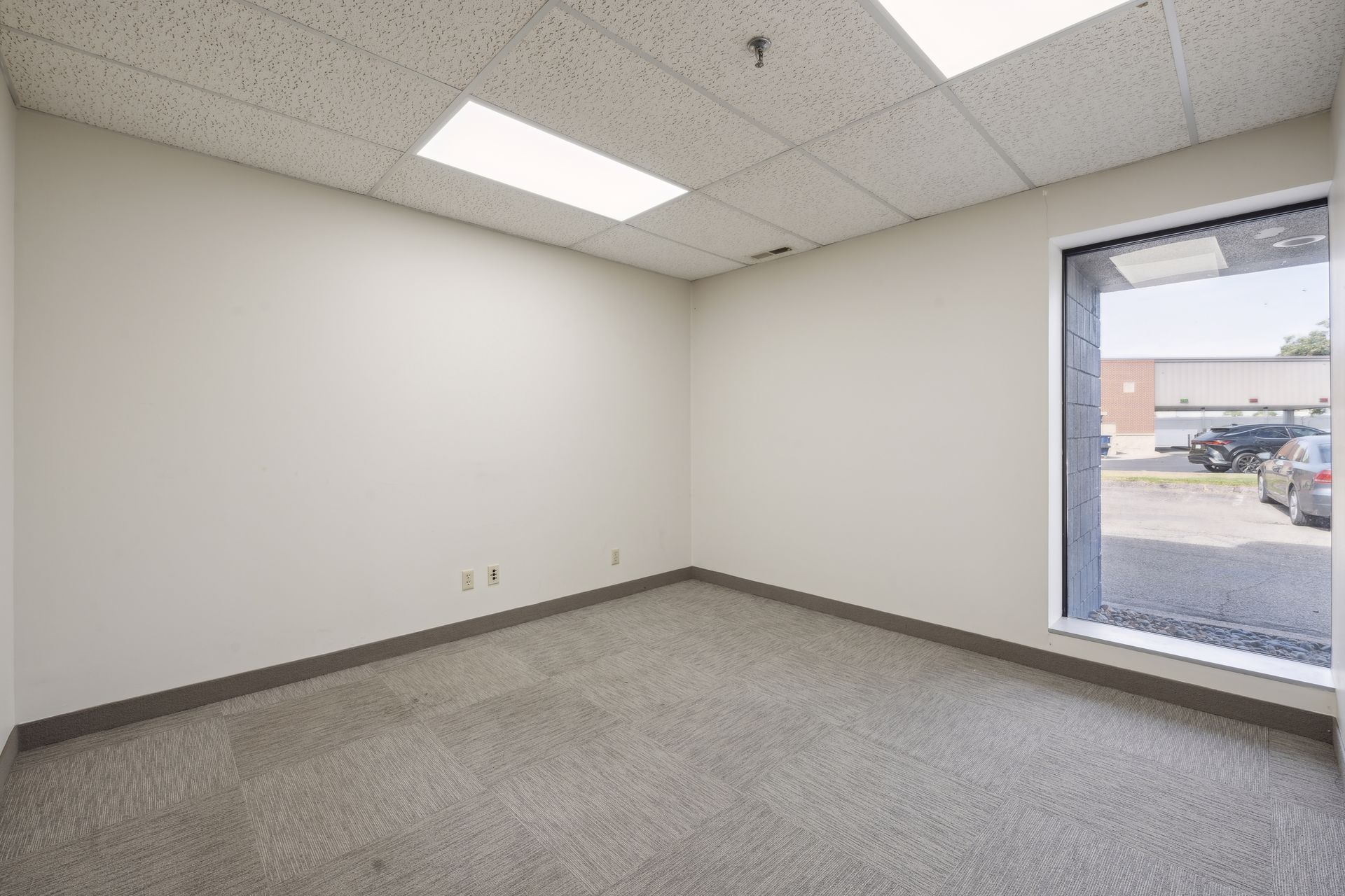 Small corner office with carpet tile flooring and one window facing the parking lot