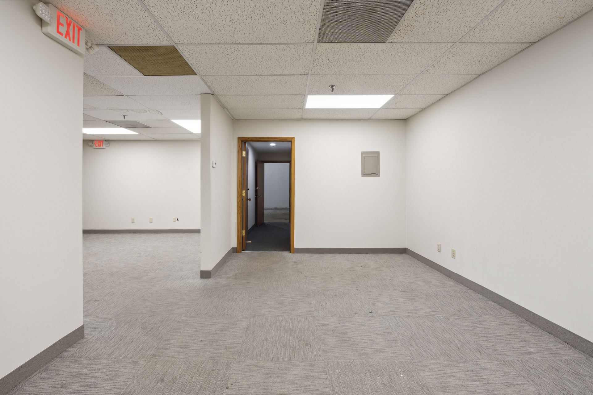 Open interior area with a suspended tile ceiling, an illuminated exit sign and a wall-mounted electrical panel
